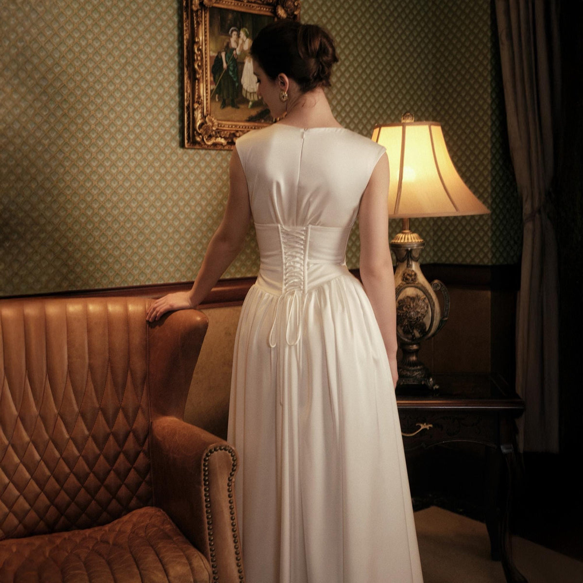 May include: A white satin wedding dress with a corset back. The dress has a fitted bodice and a flowing skirt. The model is standing near a brown leather armchair and a vintage lamp.