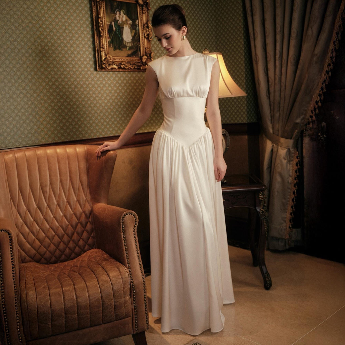 May include: A white satin gown with a fitted bodice and a flowing skirt. The dress features a high neckline and cap sleeves. The model is standing near a brown leather armchair in a room with a vintage aesthetic.