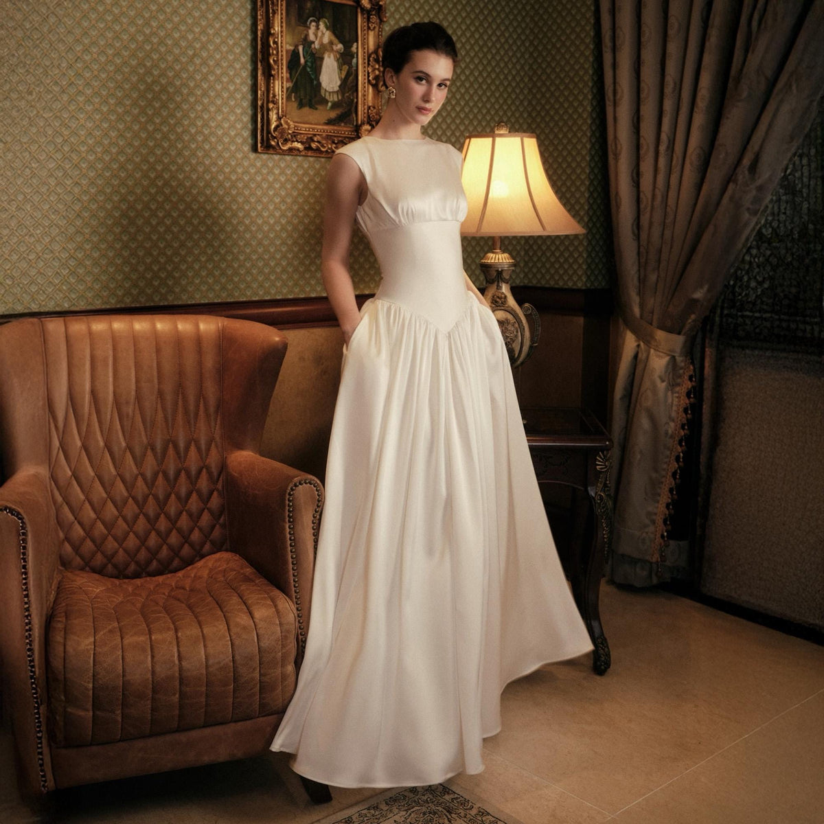 May include: A white satin wedding dress with a bateau neckline and a full skirt. The dress has a fitted bodice and pockets. The model is standing in a room with a leather armchair, a lamp, and a framed picture.
