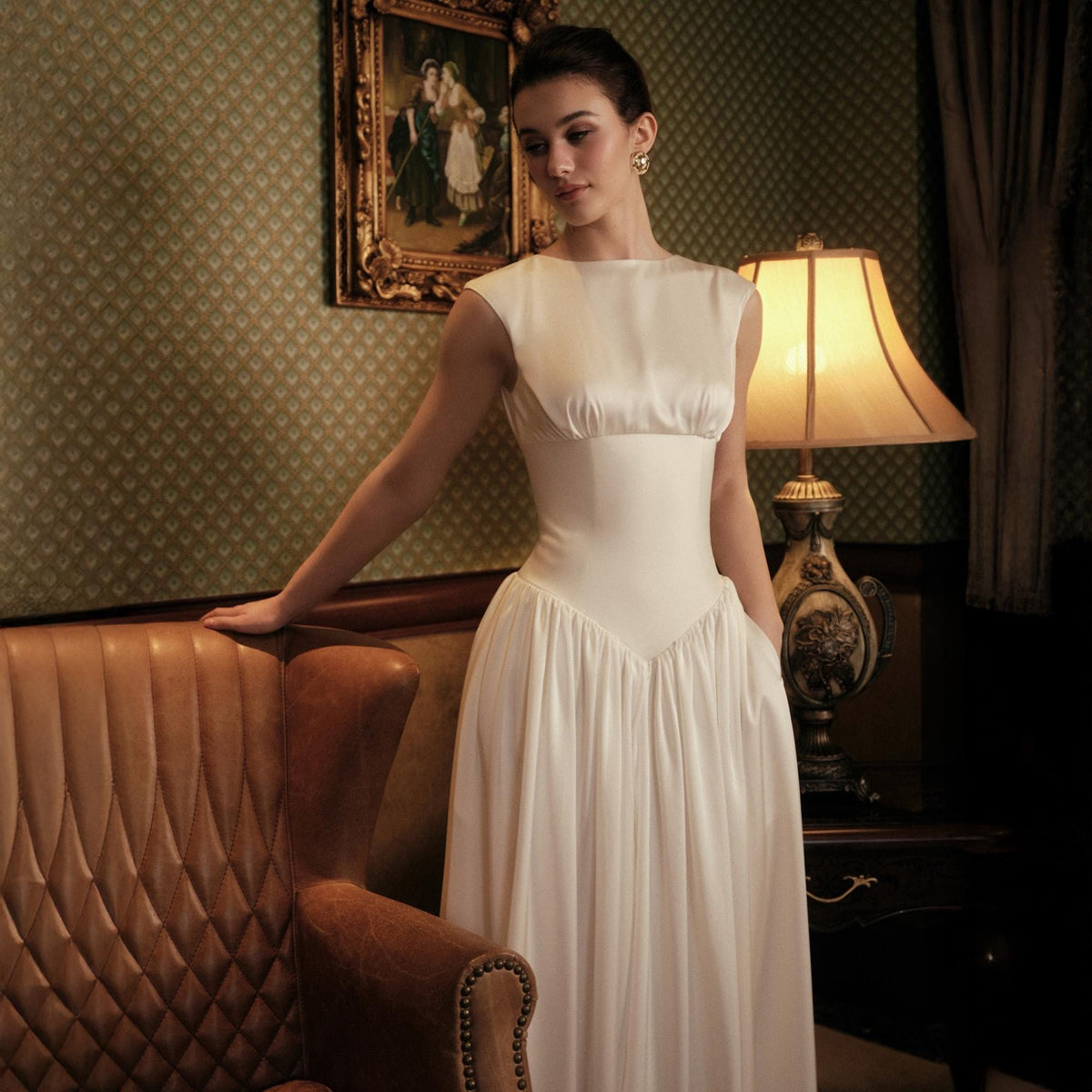 May include: A white satin dress with a fitted bodice and a flowing skirt. The dress has a scoop neckline and a defined waist. The model is standing near a brown leather armchair in a room with a vintage lamp and a framed picture on the wall.