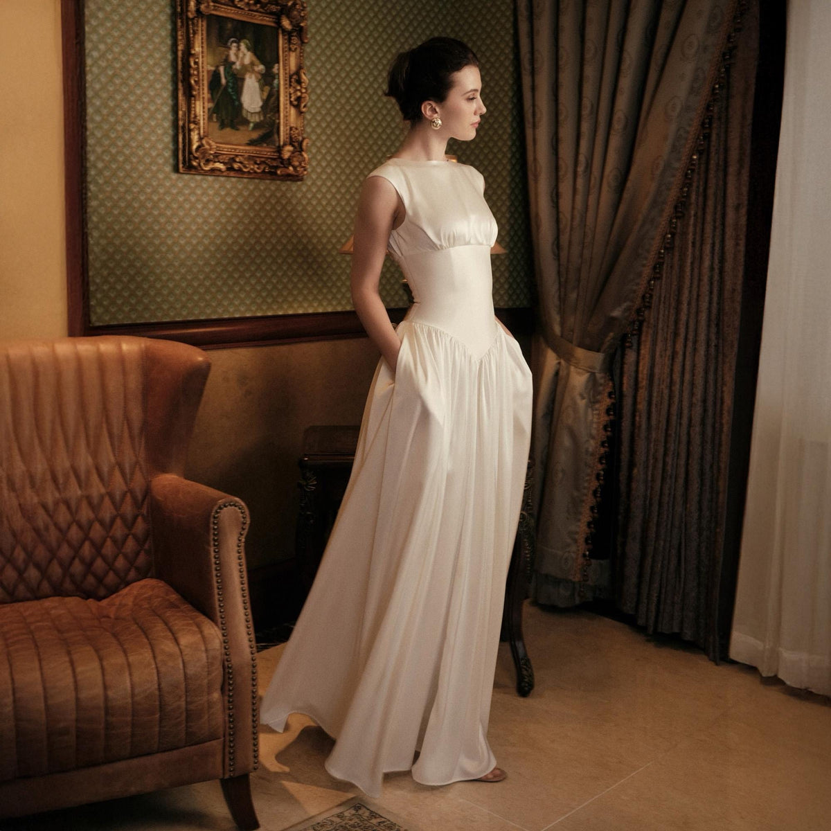 May include: A white, floor-length gown with a fitted bodice and flowing skirt. The dress features a scoop neckline and pockets. The woman is standing near a window with a brown leather armchair and a framed picture in the background.