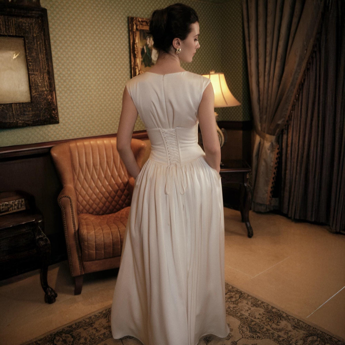 May include: A white, floor-length dress with a fitted bodice and a corset back. The dress has a simple, elegant design with a flowing skirt. The model is standing in a room with a vintage aesthetic, with a leather armchair and a patterned rug.