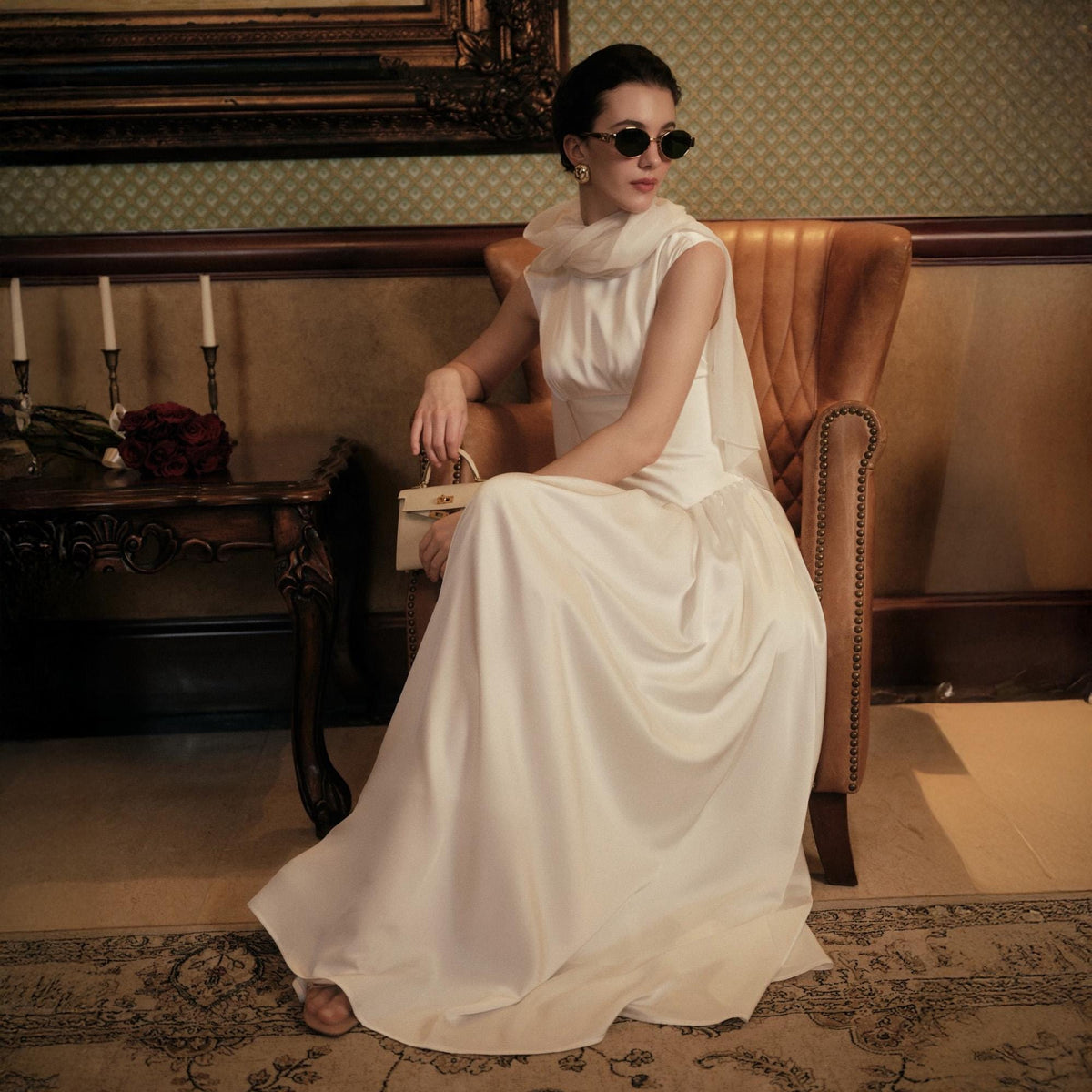 May include: A woman in a white satin gown and sunglasses sits in a leather chair. She holds a small, cream-colored handbag. The room has a vintage aesthetic, with a dark wooden side table and a patterned rug.