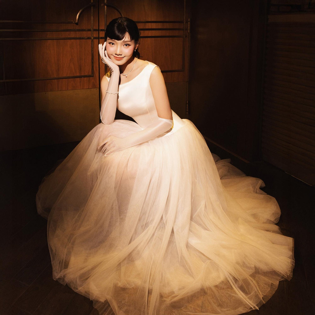 May include: A woman wearing a white sleeveless gown with a full tulle skirt and long sheer gloves. The dress has a fitted bodice and a flowing skirt. The woman is seated, with her hand resting on her cheek, and is wearing a necklace and bracelet.