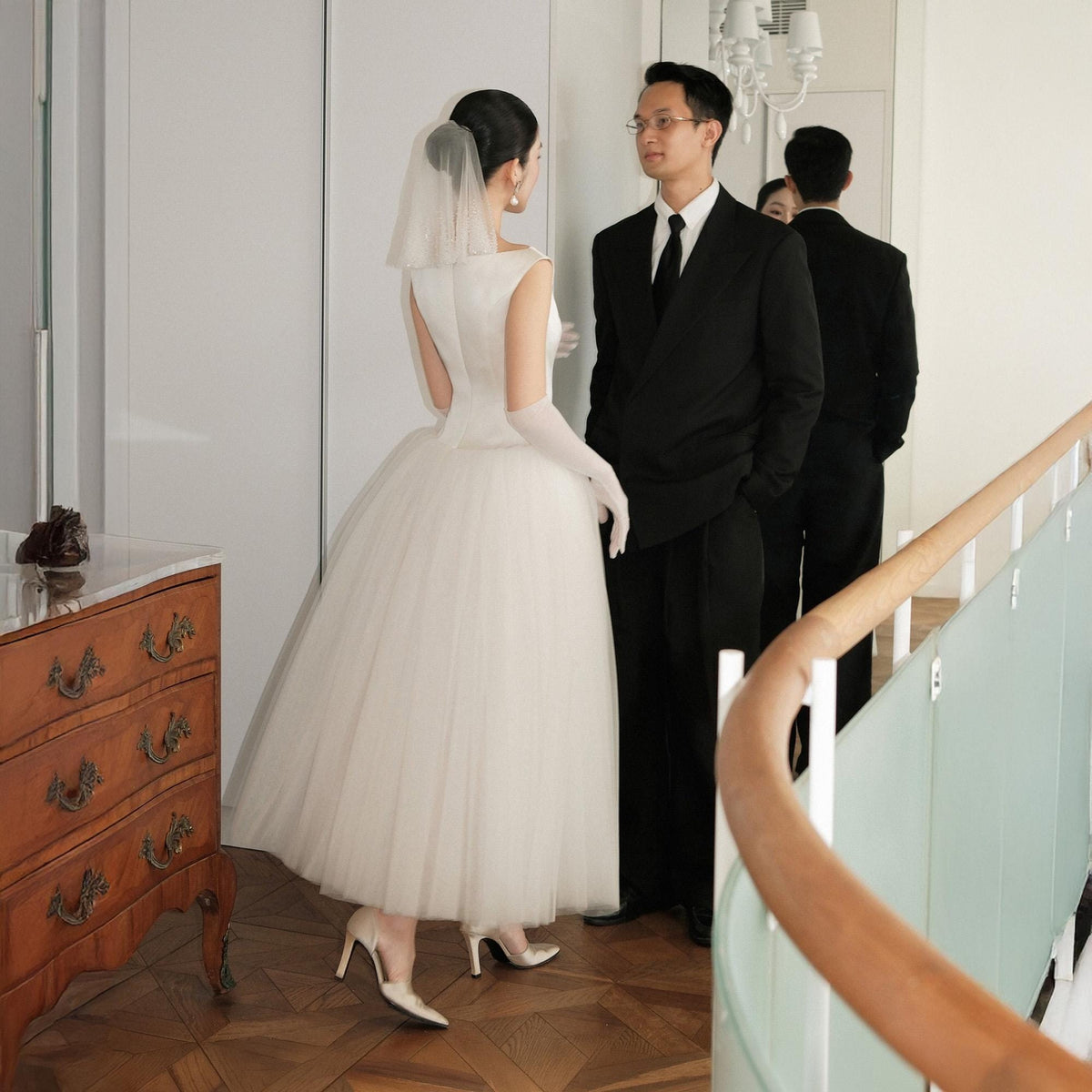 May include: A bride in a white sleeveless wedding dress with a tulle skirt and veil stands facing a groom in a black suit. The bride wears white gloves and heels. A wooden dresser is on the left.