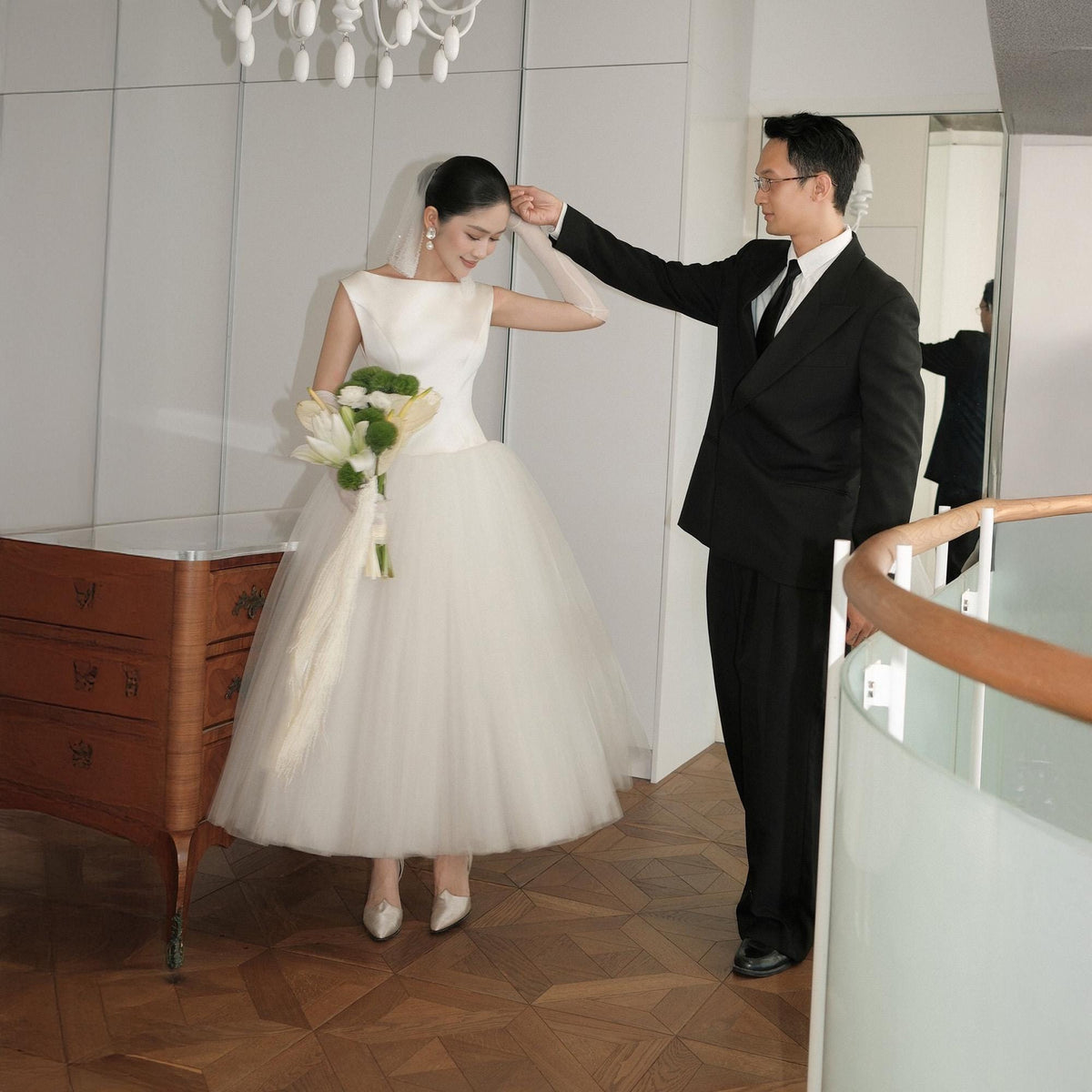 May include: A bride in a white, sleeveless wedding dress with a tulle skirt and a bouquet of white and green flowers. A groom in a black suit and tie stands beside her. The scene is indoors, with a wooden cabinet and a chandelier.