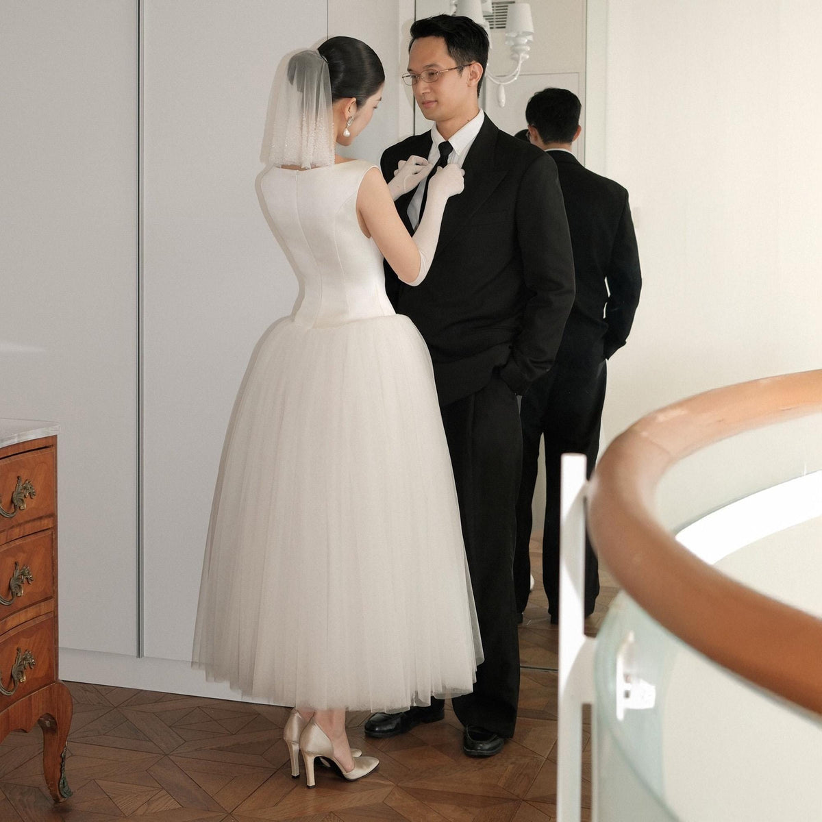 May include: A bride in a white sleeveless wedding dress with a tulle skirt and veil adjusts the groom's tie. The groom wears a black suit and glasses. A second man in a black suit stands behind them. The scene takes place indoors.