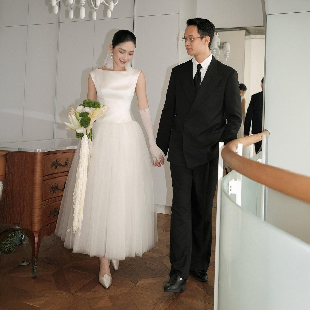 May include: A bride in a white satin and tulle wedding dress, holding a bouquet of white and green flowers, walks with a groom in a black suit. The couple is holding hands and walking on a wooden floor.