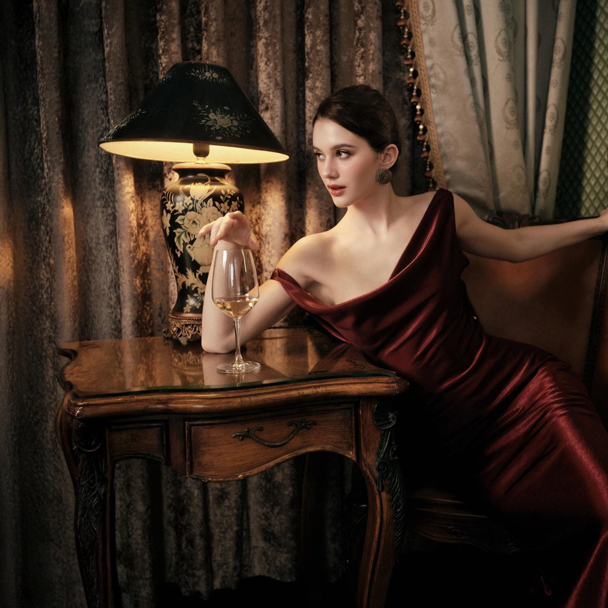 May include: A burgundy, one-shoulder evening gown. A glass of white wine sits on a dark wood table next to a decorative lamp. The lamp has a black shade and a floral design on the base. The scene is set in a room with dark curtains.