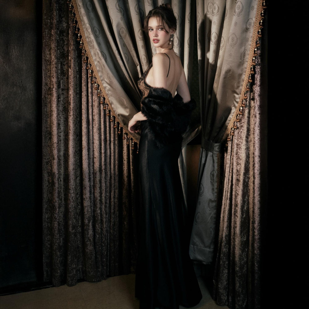 May include: A woman wearing a black evening gown with spaghetti straps and a fur stole. The dress has a low back and a flowing skirt. The woman is standing in front of a dark curtain.