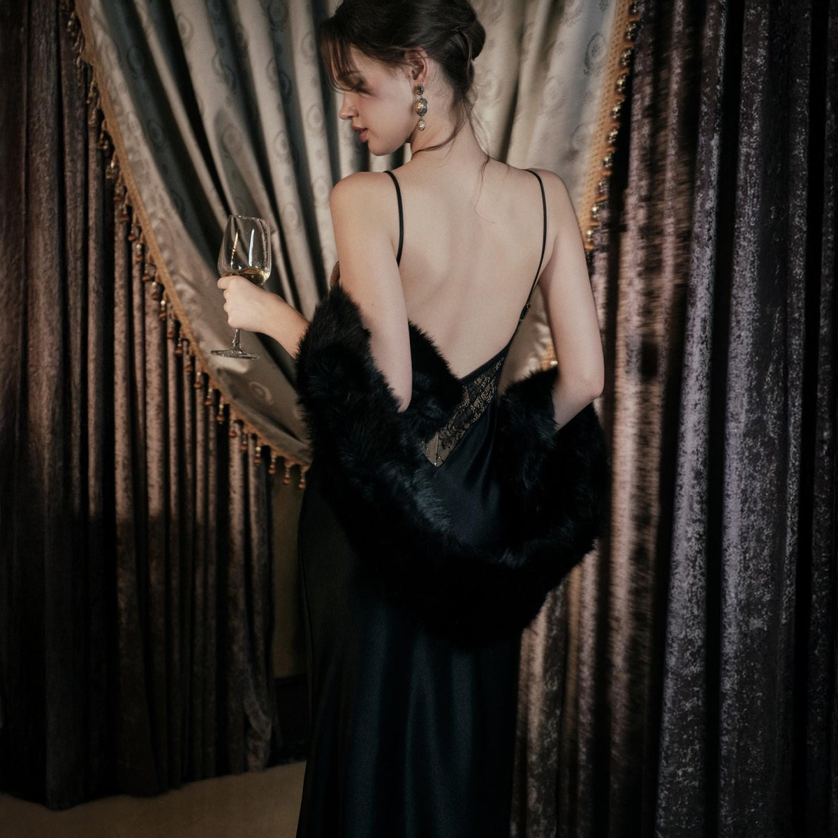 May include: A black satin slip dress with spaghetti straps and a low back. A black faux fur wrap is draped over the shoulders. The woman is holding a wine glass. The background features a dark curtain.