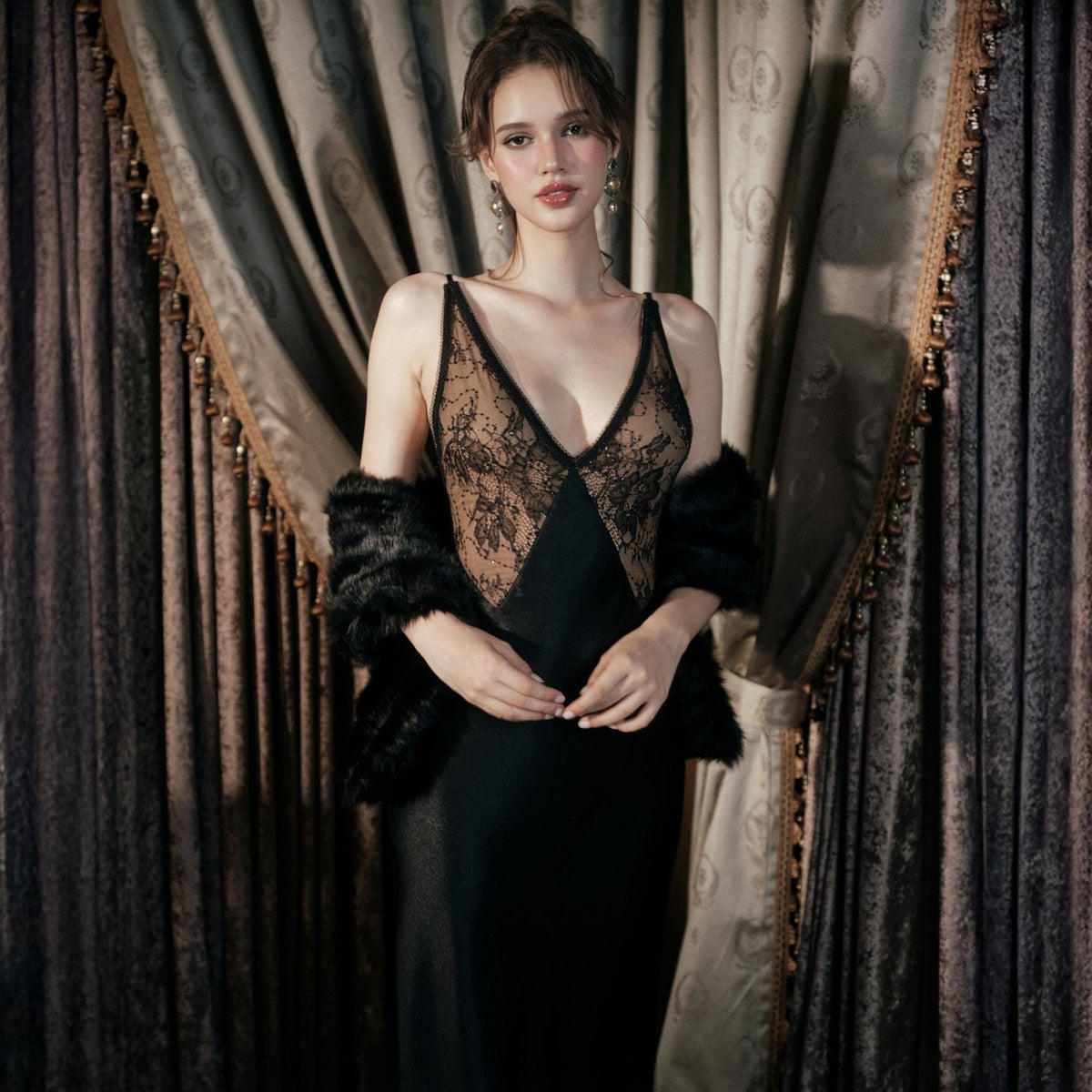 May include: A black slip dress with a V-neckline and a lace bodice. A black faux fur wrap is draped over the shoulders. The dress is floor-length and made of a silky material. The background features ornate curtains.