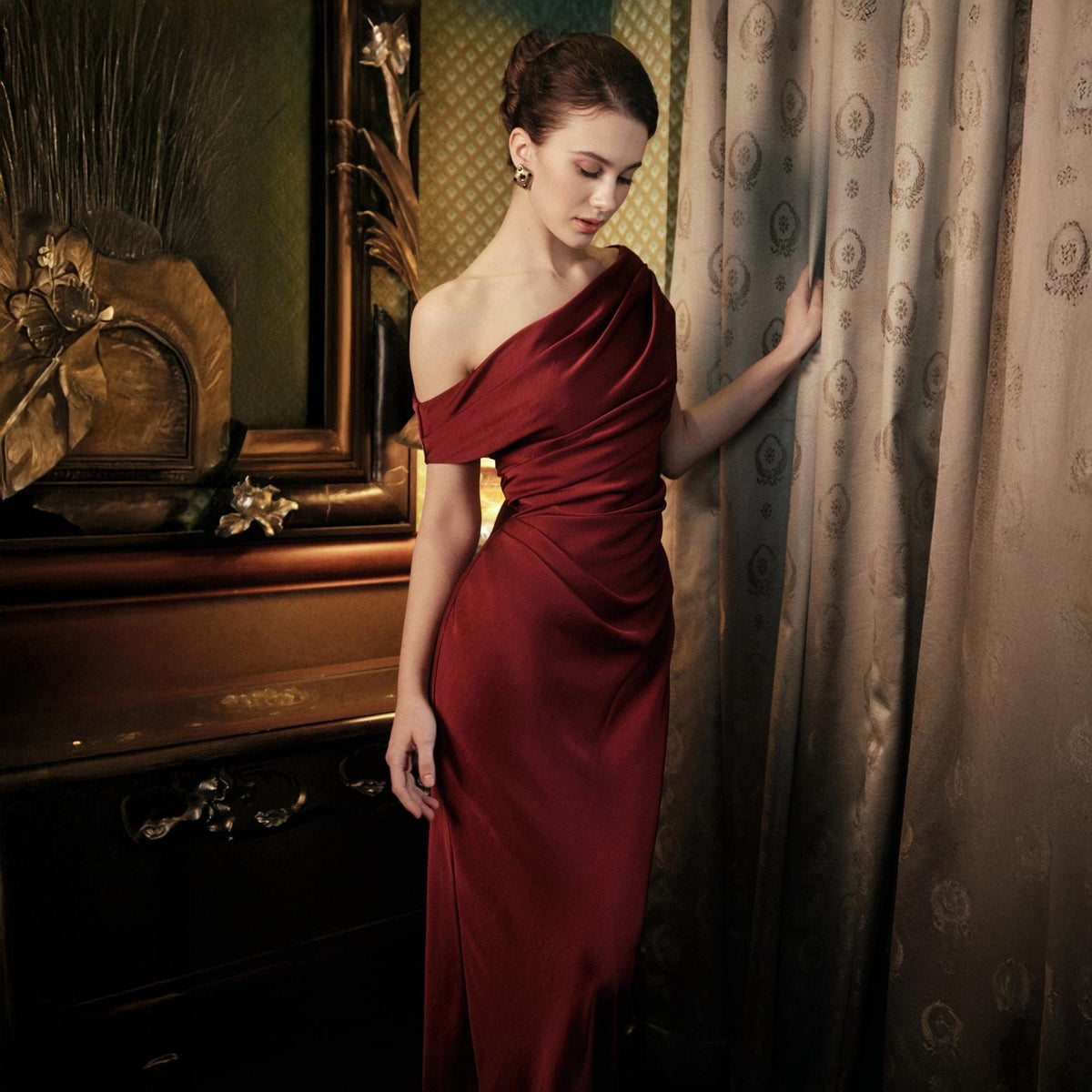 May include: A formal, one-shoulder, floor-length gown in a deep red hue. The dress features draped fabric across the bodice and a sleek, fitted skirt. The model is standing near a patterned curtain and a dark wooden frame.