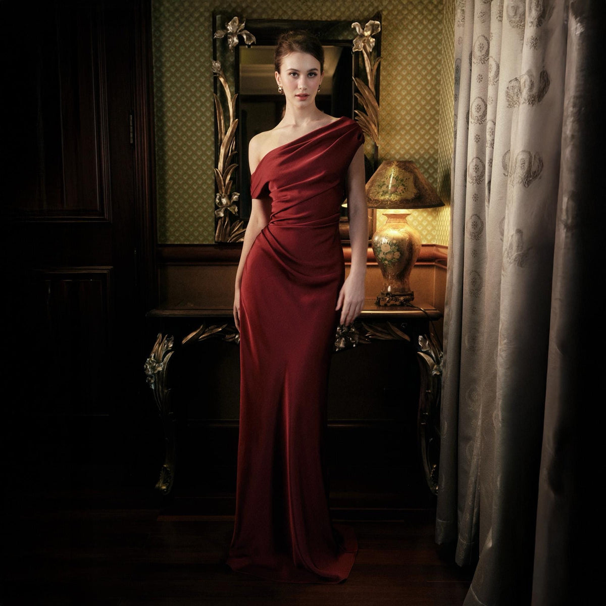 May include: A floor-length, one-shoulder, burgundy-colored gown. The dress features a draped bodice and a fitted silhouette. The woman is standing in a room with a mirror, a table, and a lamp.