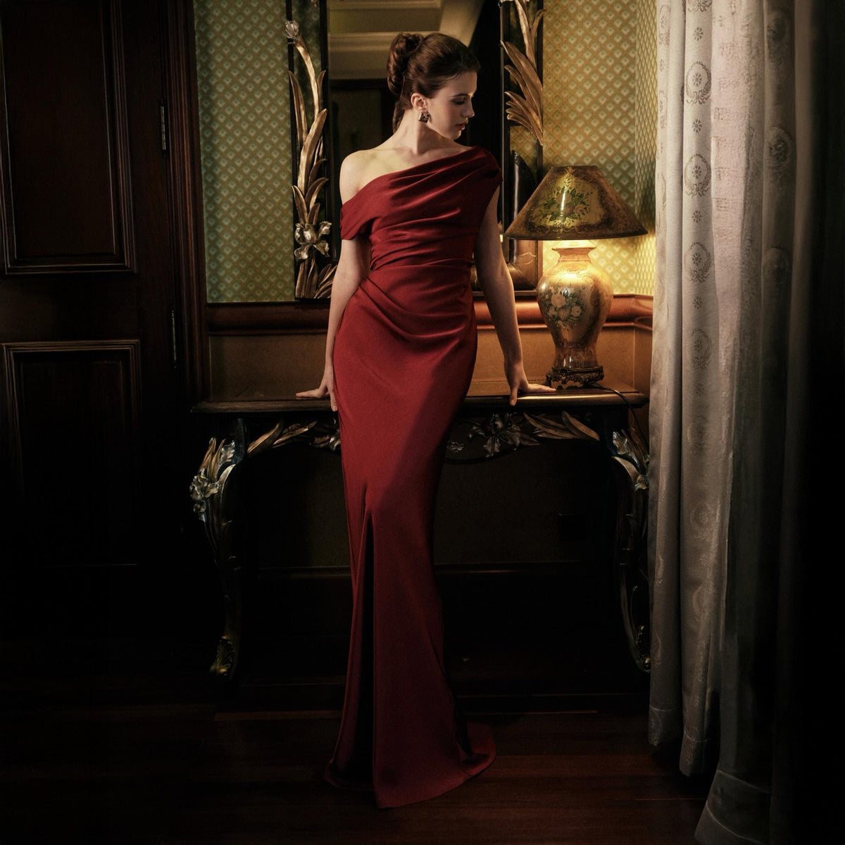 May include: A floor-length, one-shoulder, crimson red gown. The dress features a draped neckline and a fitted silhouette. The image is set in a dimly lit room with a decorative mirror and a vintage lamp.