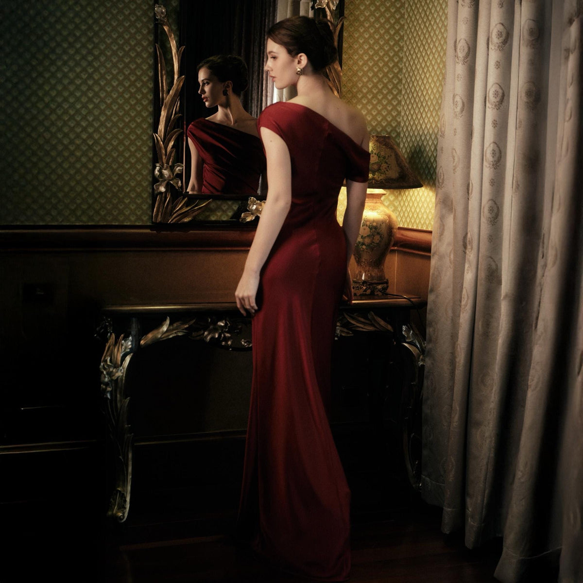 May include: A long, flowing, one-shoulder, dark red gown. The dress is made of a silky material and has a subtle sheen. The image is taken in a room with a mirror and a decorative table. The dress is the focal point.