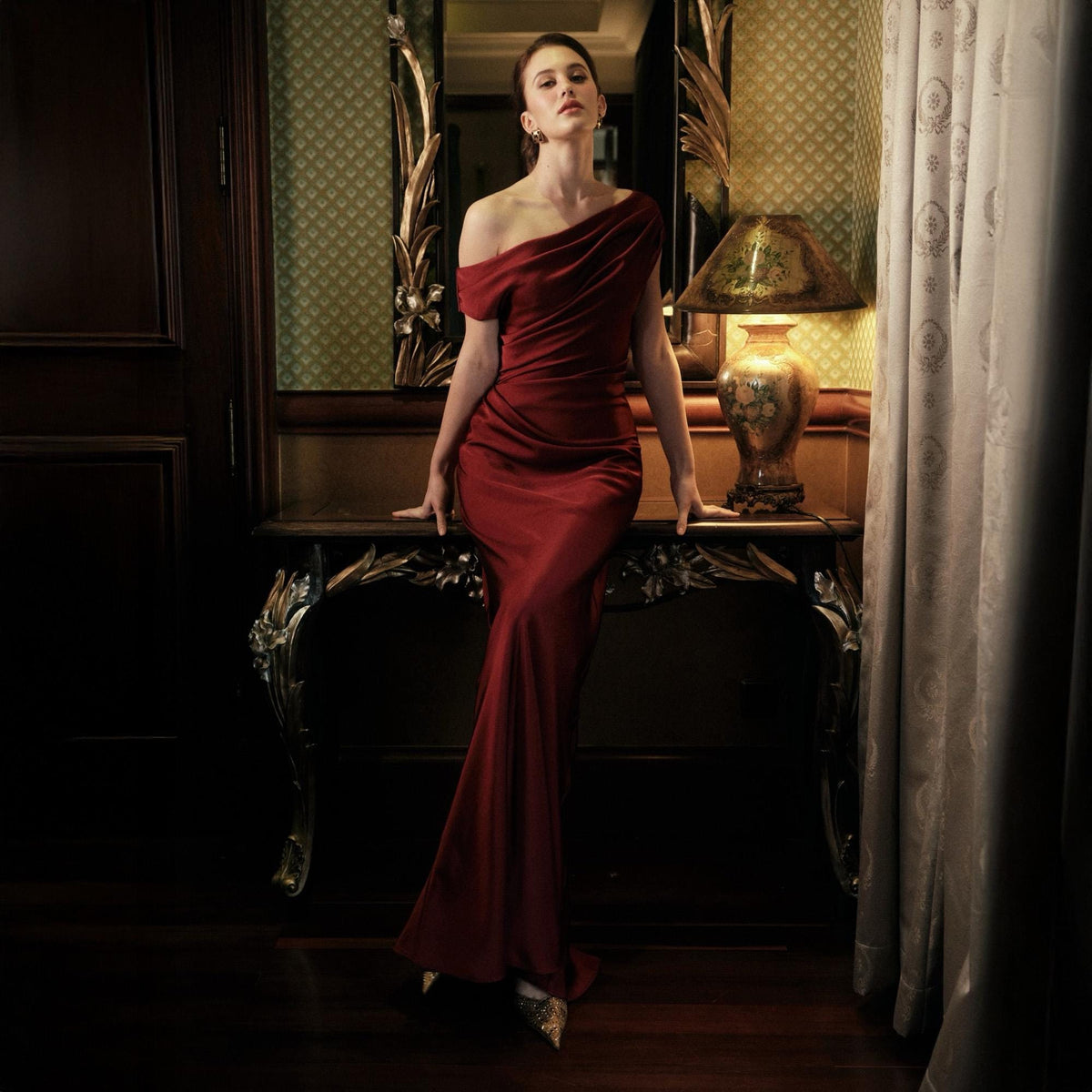 May include: A floor-length, one-shoulder, crimson red gown. The dress features a draped bodice and a fitted silhouette that flares out at the hem. The image is set in a room with a decorative mirror and a vintage lamp.