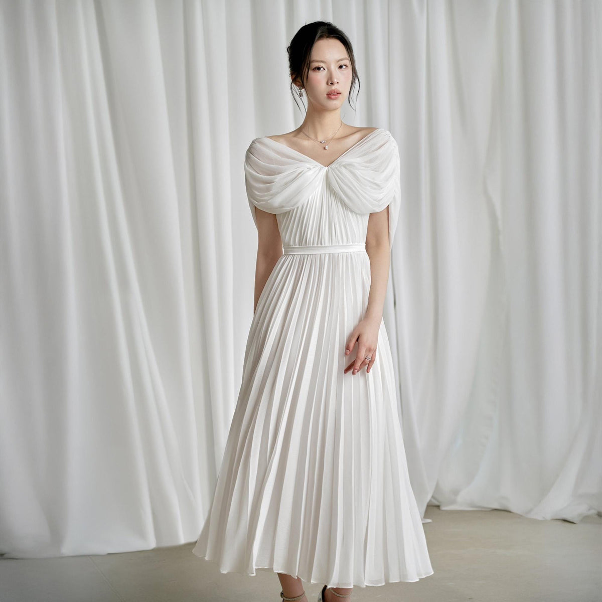 May include: A white, off-the-shoulder dress with a pleated skirt. The dress features a draped bodice and a thin waist belt. The fabric appears to be lightweight and flowing, suitable for formal occasions. The dress is knee-length.