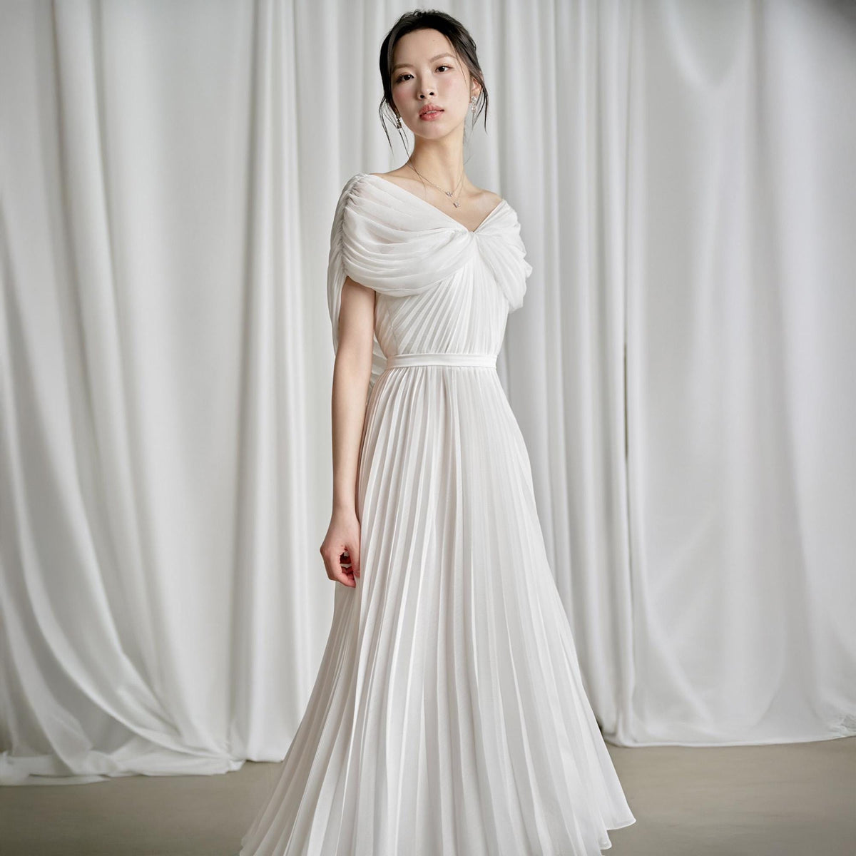 May include: A white pleated gown with a draped bodice and a fitted waist. The dress features a flowing skirt and a delicate neckline. The model is wearing a necklace and earrings. The background is a white curtain.