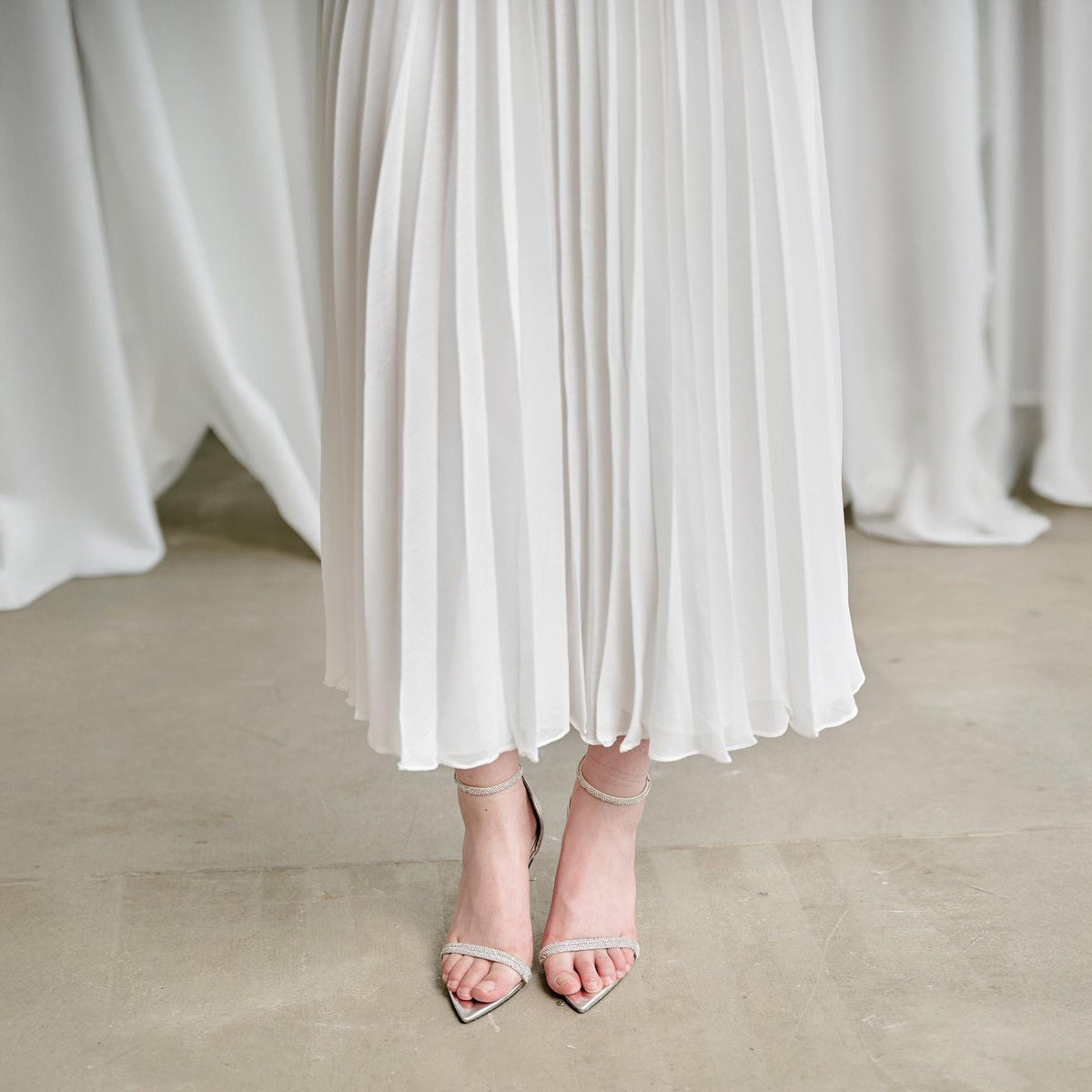 May include: A pair of silver high-heeled sandals with thin straps and pointed toes. The sandals are adorned with small, sparkling embellishments. The person is wearing a white pleated skirt that falls to the ankles.