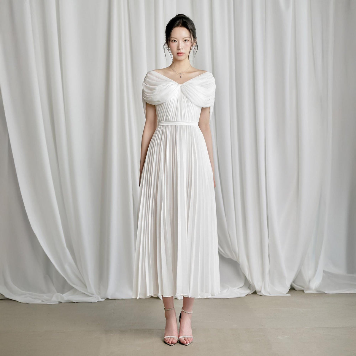 May include: A white pleated midi dress with a draped neckline and a fitted waist. The dress has a flowing skirt and is paired with strappy heels. The background features white curtains.
