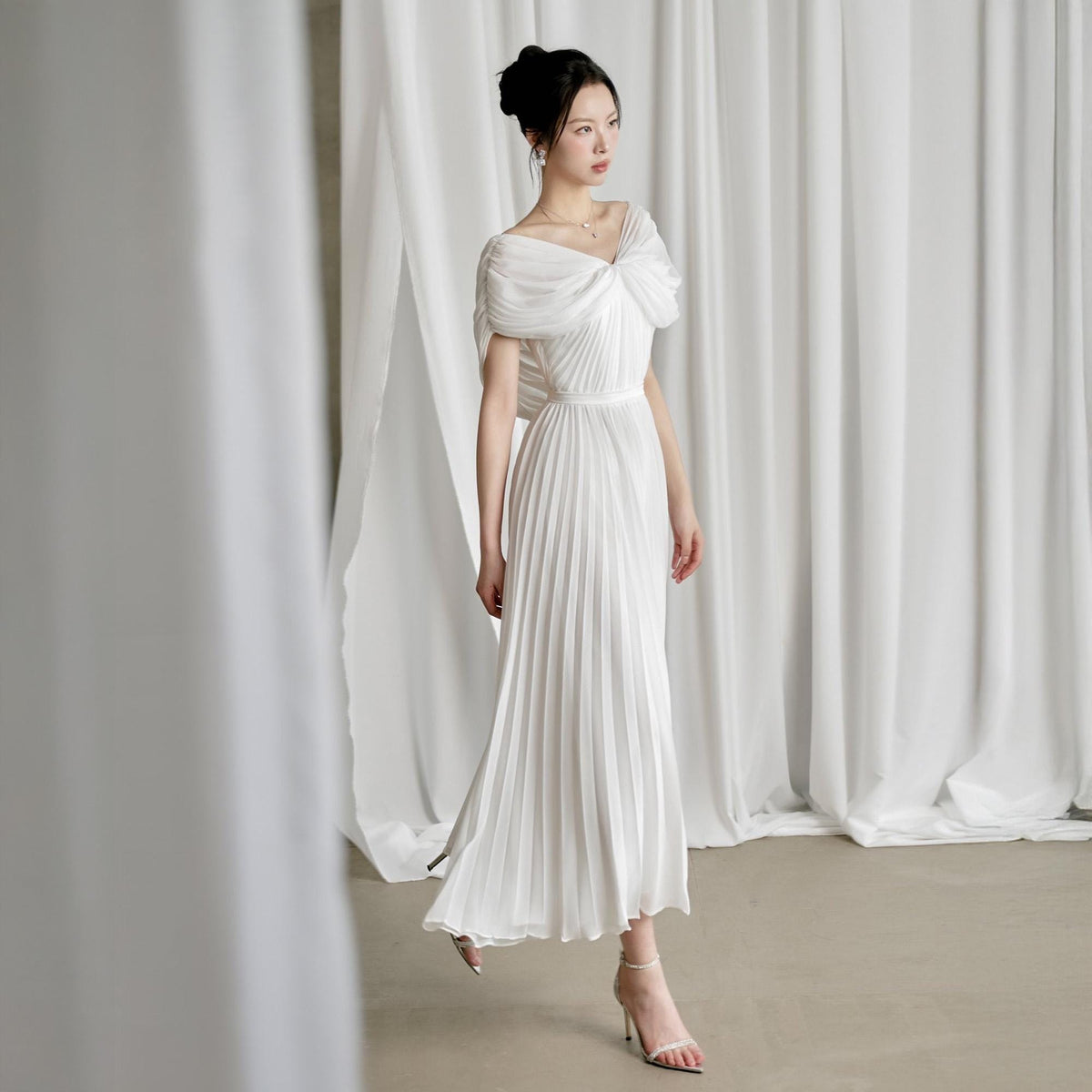 May include: A white, floor-length dress with a pleated skirt and off-the-shoulder design. The dress features a fitted waist and a draped bodice. The model is wearing silver high-heeled sandals. The background is a white curtain.