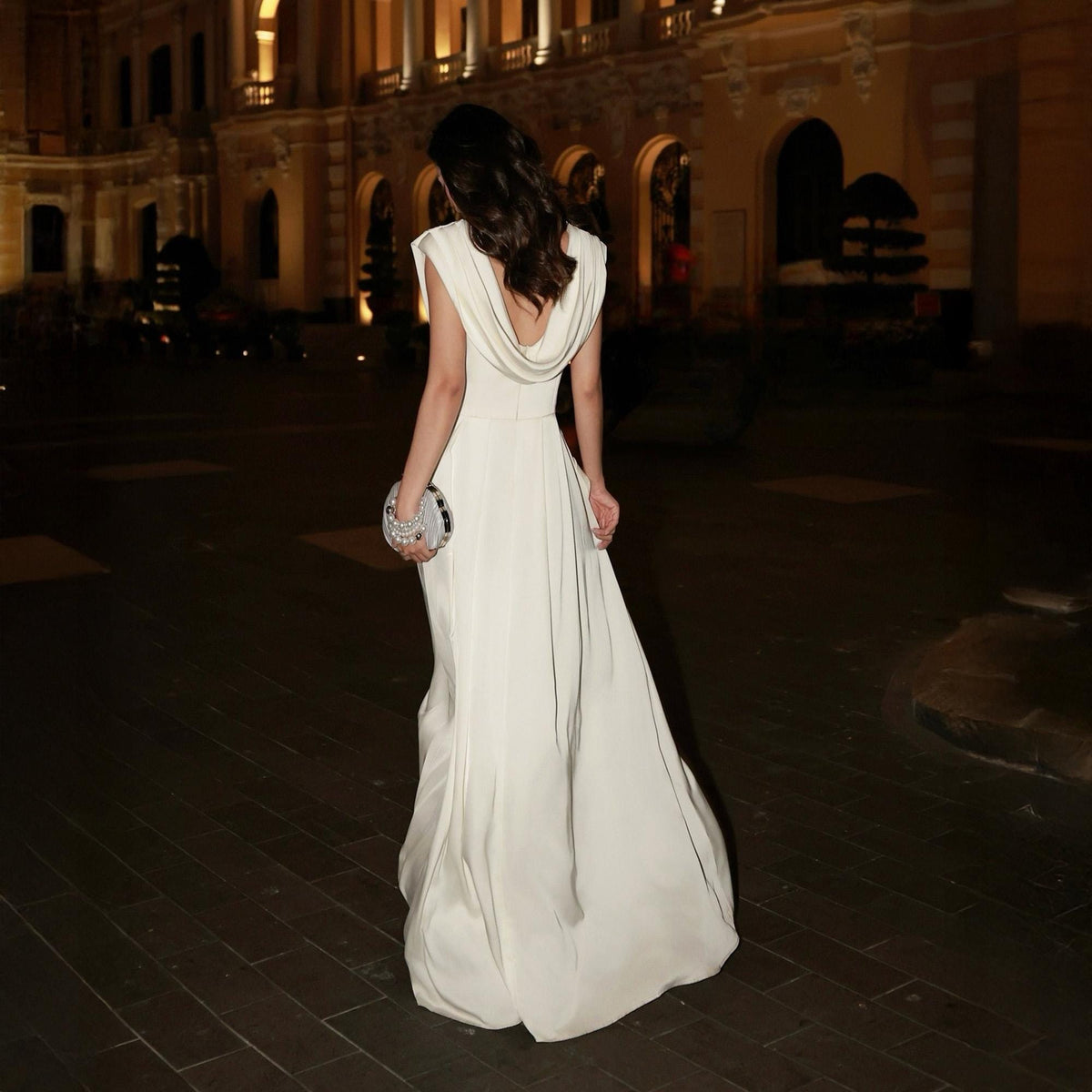 May include: A long, ivory-colored gown with a draped cowl neckline and a flowing skirt. The dress is paired with a small, silver clutch. The image is taken at night, with a blurred background of a building.