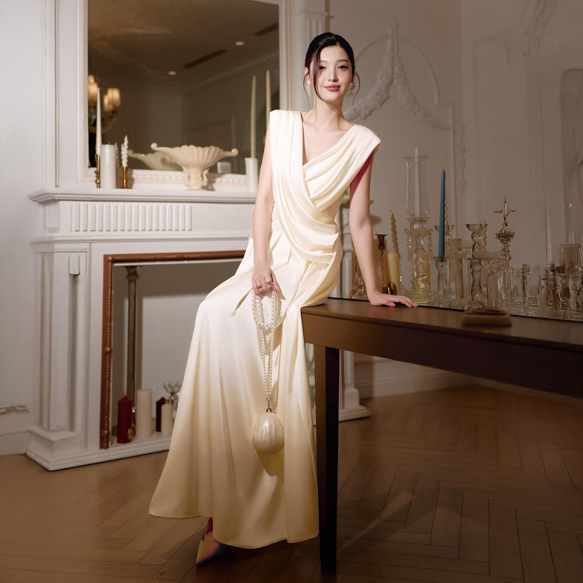 May include: A cream-colored, draped evening gown with a V-neckline and a flowing skirt. The dress features a subtle red accent at the neckline. A pearl necklace and a matching clutch complete the elegant look. The setting is a room with a fireplace and a dark wood table.