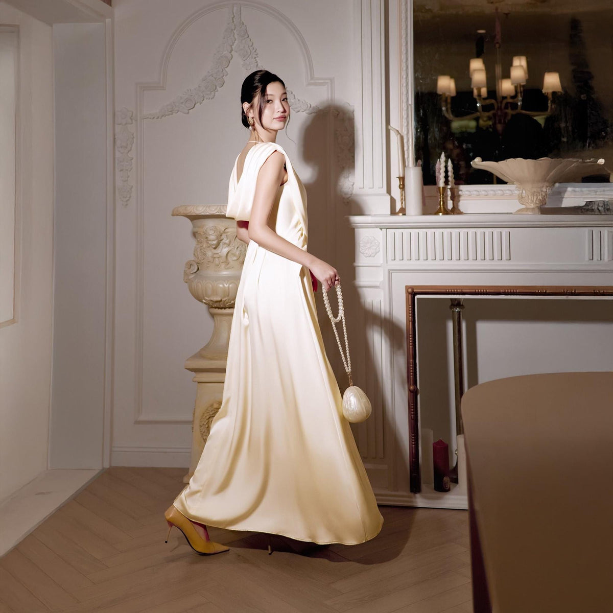 May include: A woman in a long, flowing, cream-colored satin gown with a matching purse and pearl necklace. She is wearing yellow heels. The dress has a draped neckline and a subtle train. The background includes a fireplace and ornate architectural details.