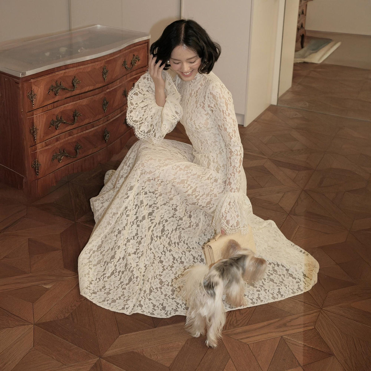 May include: A floor-length cream-colored lace dress featuring a high neckline, long sleeves with flared cuffs, and an intricate floral pattern throughout the fabric. The garment is worn with a small, light-colored clutch bag held in one hand.