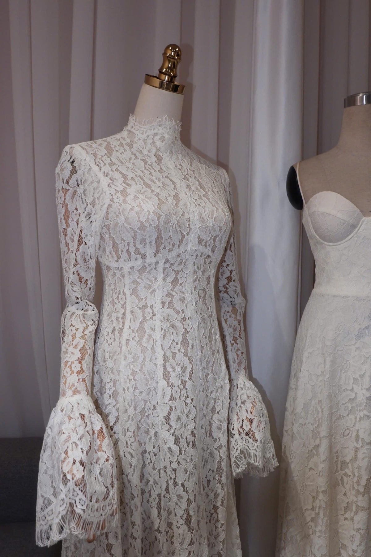 May include: A long-sleeved white lace dress featuring a high mock neckline and flared bell sleeves with fringed edges. The dress is displayed on a mannequin, showcasing the intricate floral lace pattern throughout the fitted bodice and sleeves.
