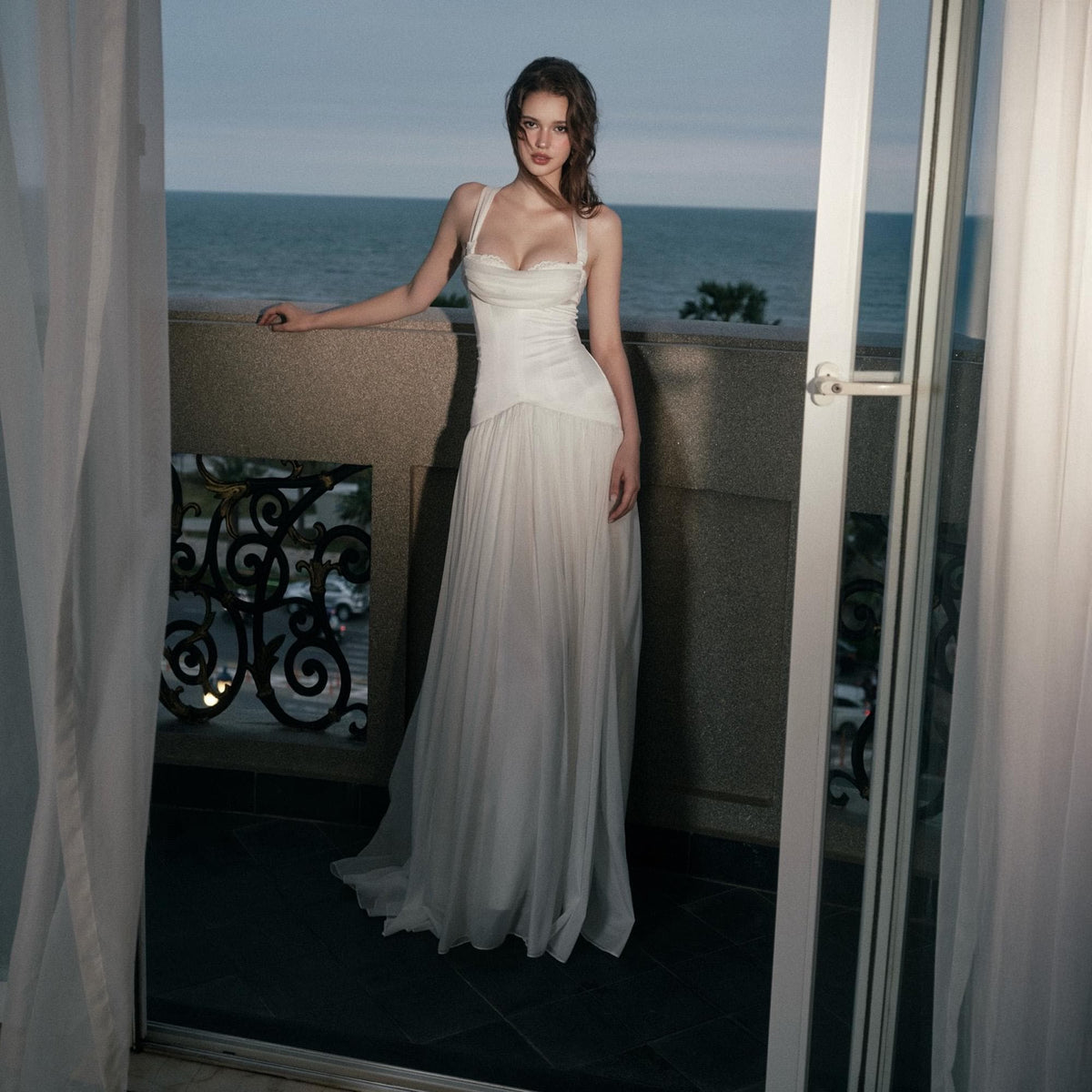 May include: A floor-length white gown featuring a fitted bodice with a sweetheart neckline and thick straps, transitioning into a flowing, pleated skirt. The garment is constructed with a smooth, structured upper section and a sheer, layered lower skirt.