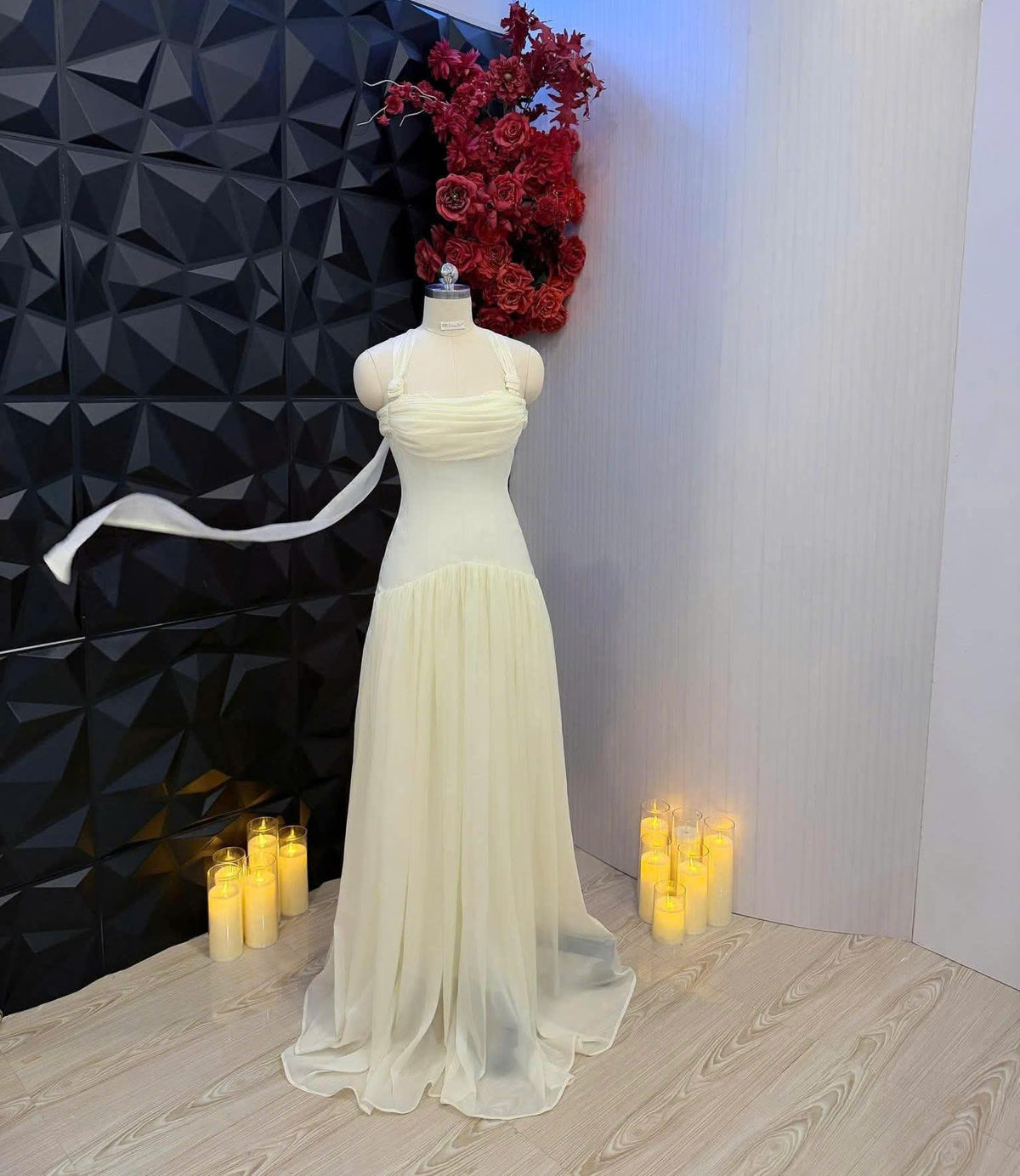 May include: A floor-length, pale yellow sleeveless dress on a mannequin, featuring a ruched bodice, a halter-style neckline with thin straps, and a flowing, sheer layered skirt. A long, thin fabric tie extends from the side of the bodice.