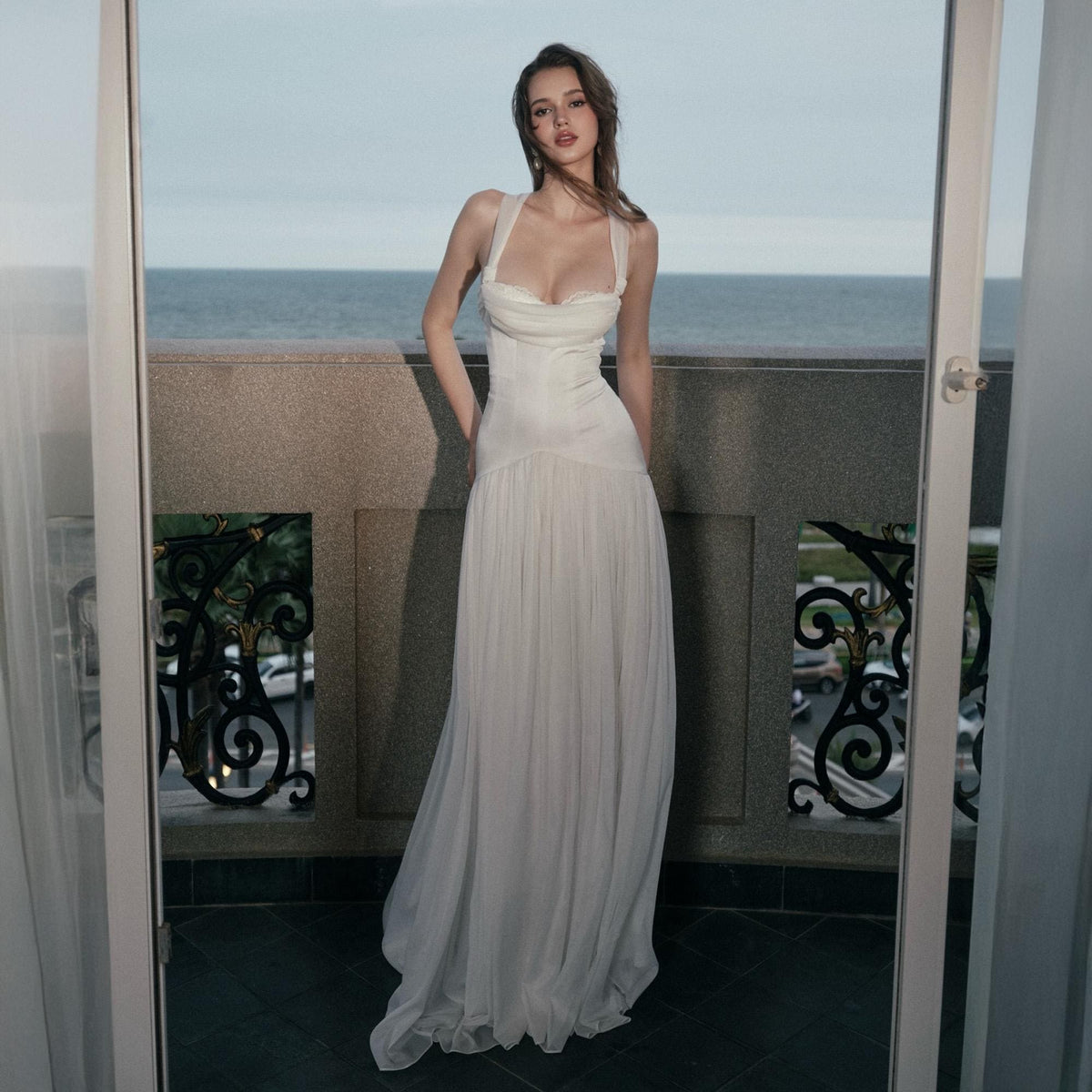 May include: A floor-length white gown featuring a draped bodice with a sweetheart neckline and thick shoulder straps. The dress has a fitted waistline that transitions into a long, flowing, pleated skirt that reaches the floor in a straight silhouette.
