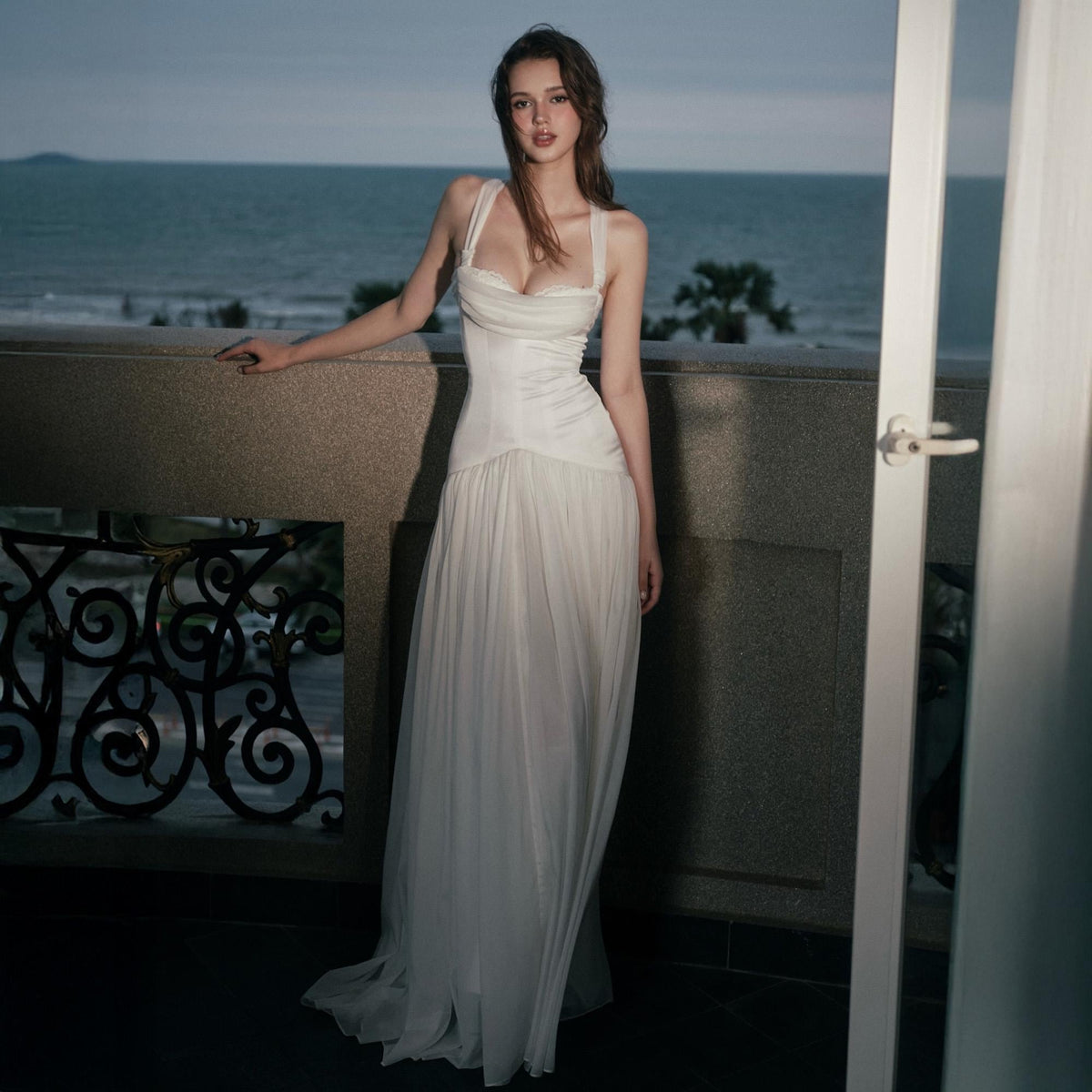 May include: A floor-length white gown featuring a structured bodice with a draped neckline and thick straps. The waistline is fitted, transitioning into a flowing, pleated skirt made of a sheer, lightweight fabric that reaches the floor.