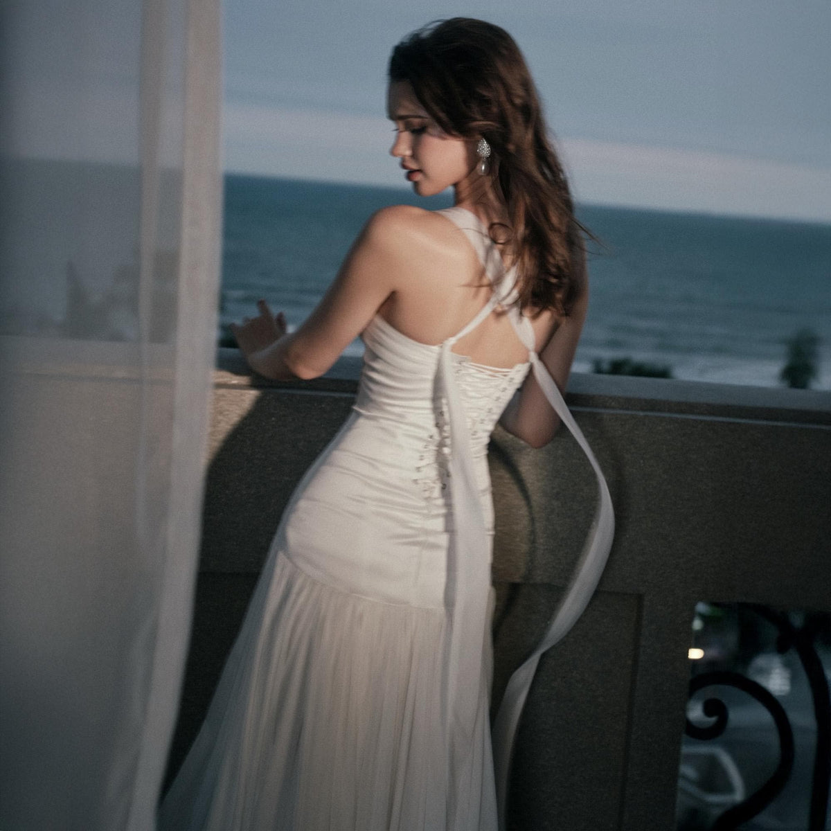 May include: A white floor-length dress featuring a fitted bodice with a lace-up back closure, thin shoulder straps, and a flowing skirt. The garment is constructed with a smooth, opaque fabric on the upper section and a sheer, pleated fabric on the lower portion.