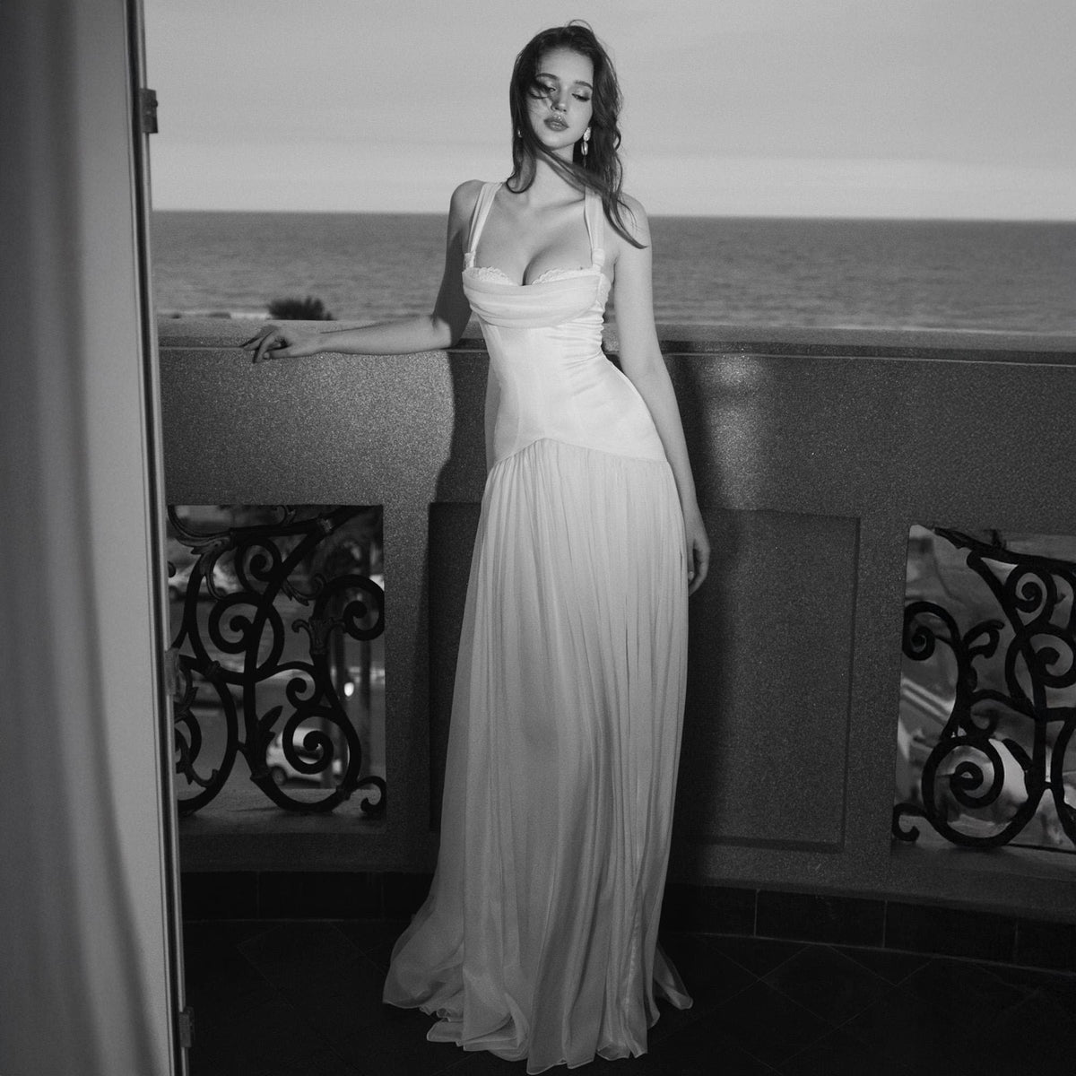 May include: A floor-length white gown featuring a fitted bodice with a draped sweetheart neckline, thick shoulder straps, and a flowing, pleated skirt that extends to the floor. The garment is captured in a black and white photograph.