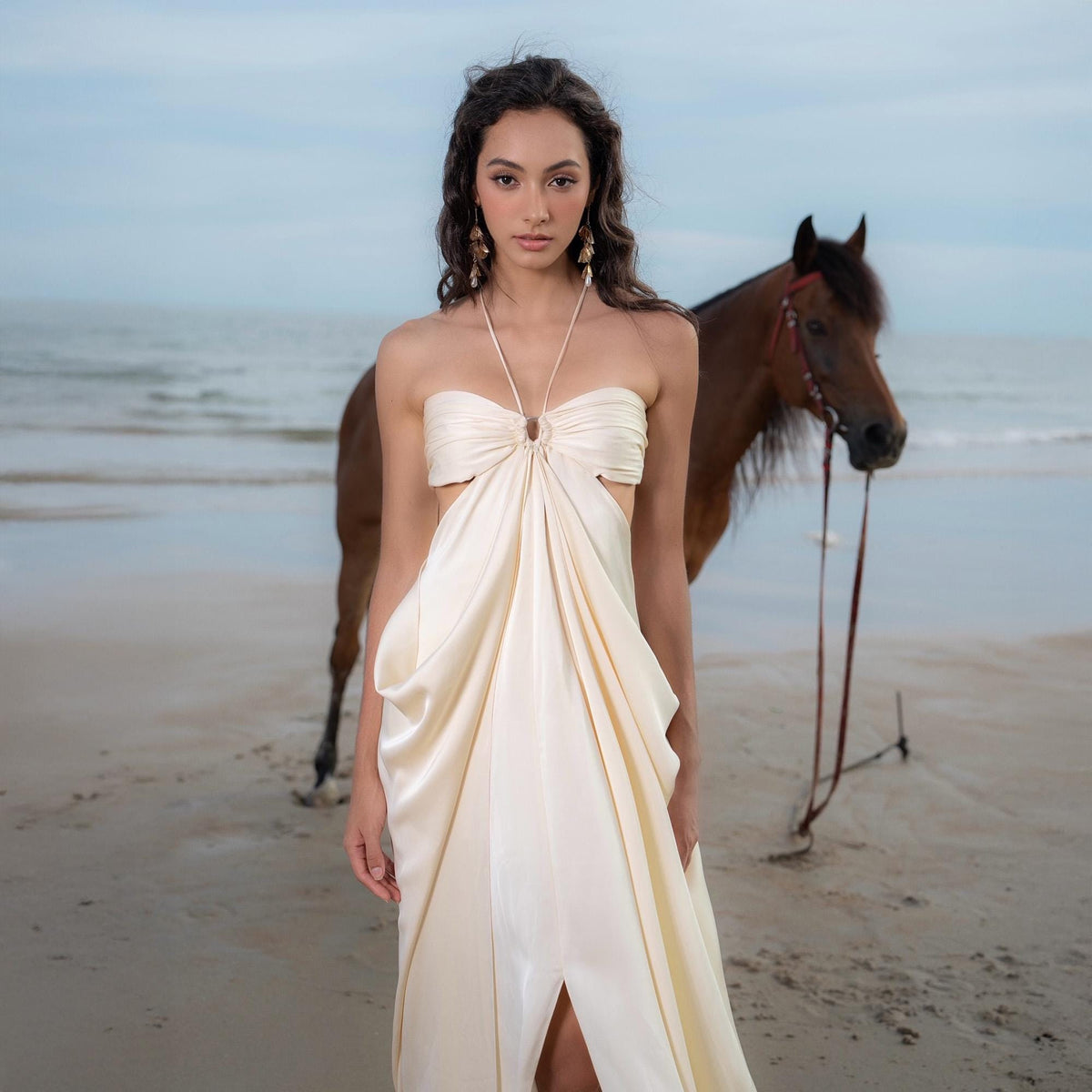 May include: A cream-colored, strapless dress with a halter neck and a front slit. The dress features cut-out details and a draped design. A brown horse stands in the background on a sandy beach.