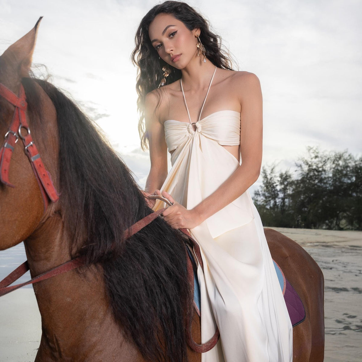 May include: A woman in a cream-colored halter dress with a keyhole cutout, riding a brown horse. The dress features a ring detail at the bust and a flowing skirt. The horse has a dark mane and is wearing a red bridle. The background is a beach scene.
