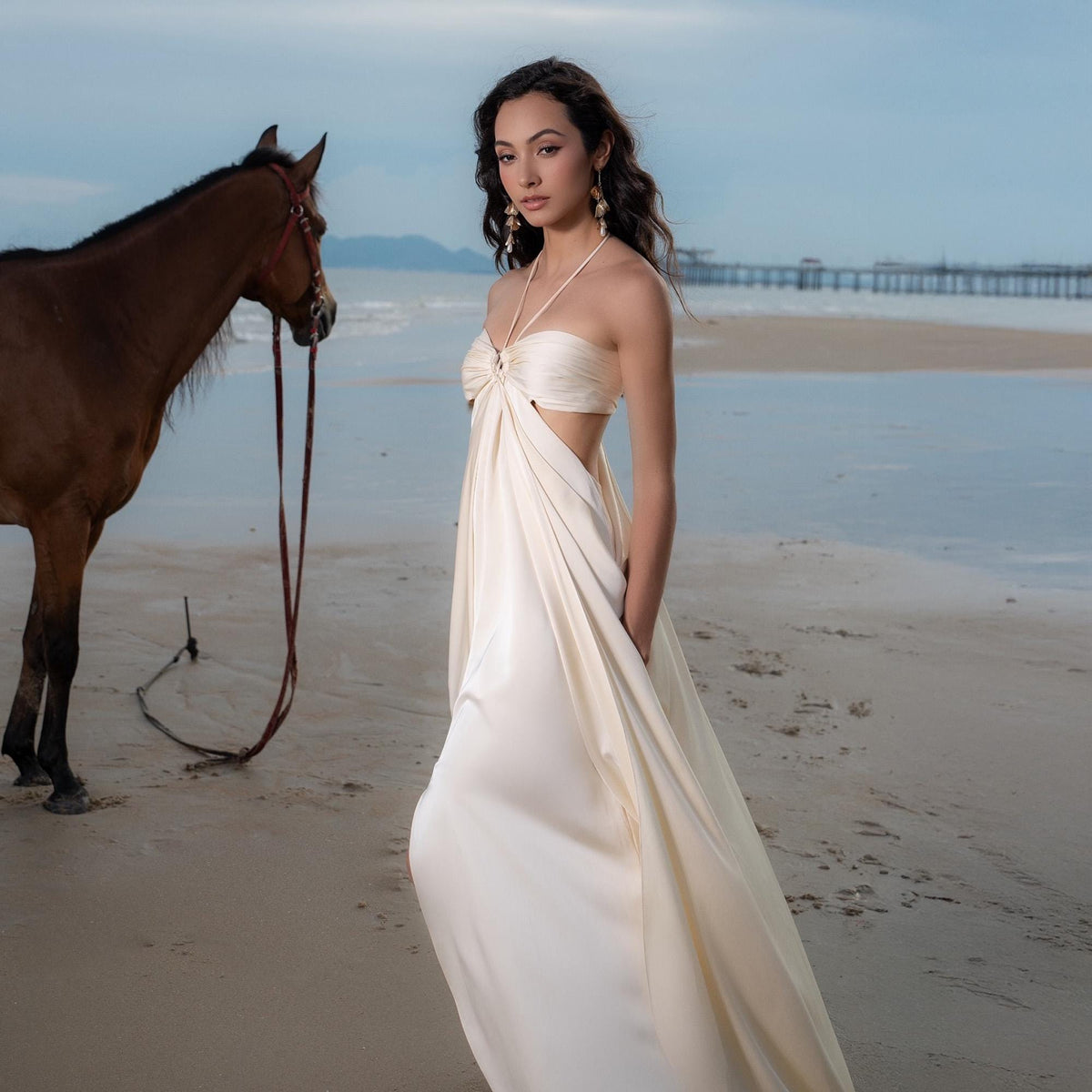May include: A woman in a cream-colored halter dress with a flowing skirt stands on a sandy beach. A brown horse with a bridle and reins is to the left. The background includes the ocean, a pier, and a cloudy sky.