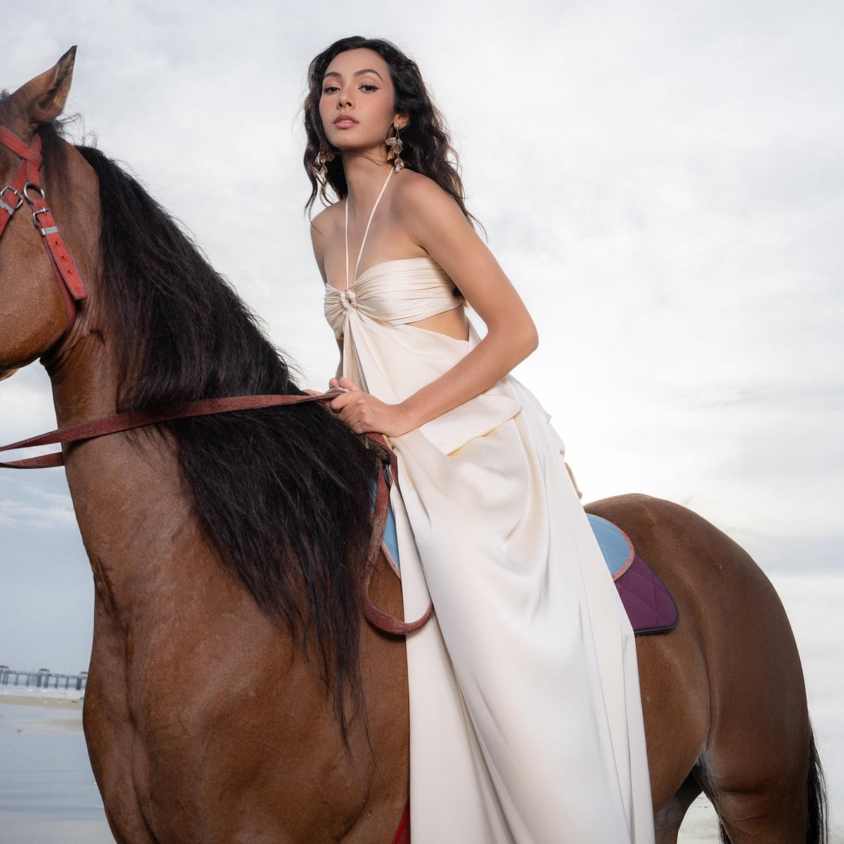 May include: A woman in a cream-colored halter dress sits atop a brown horse with a dark mane. The horse wears a red bridle and is saddled with a blue and purple saddle. The background is a cloudy sky and a beach.