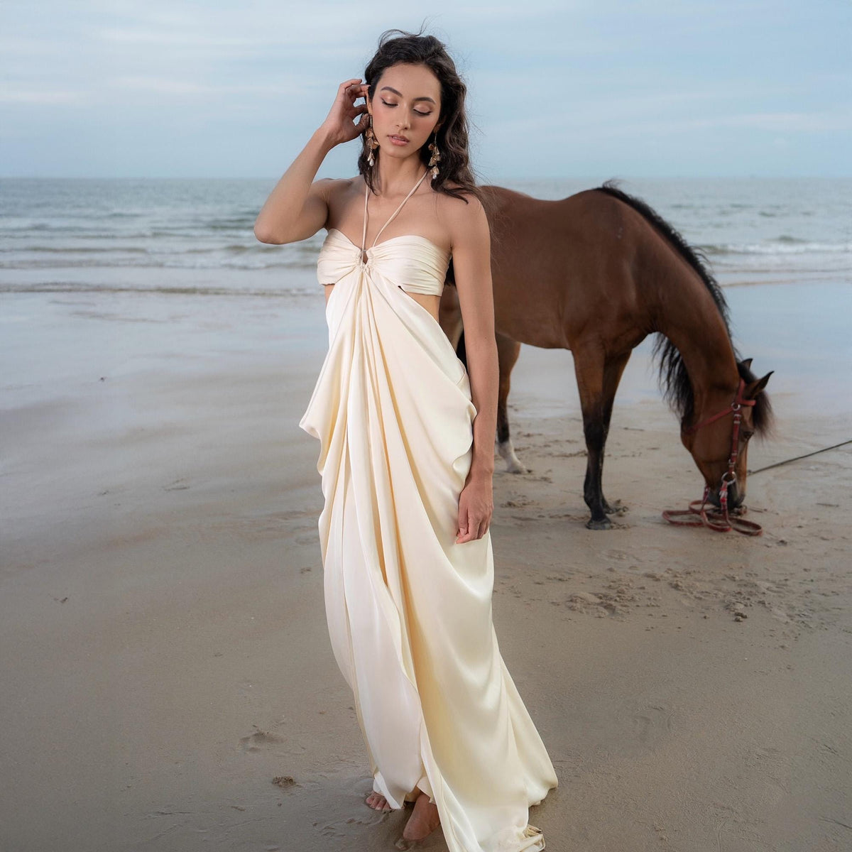 May include: A woman wearing a cream-colored, draped silk dress with a halter neckline stands on a sandy beach. A brown horse grazes in the background near the ocean. The dress has a flowing, elegant design.