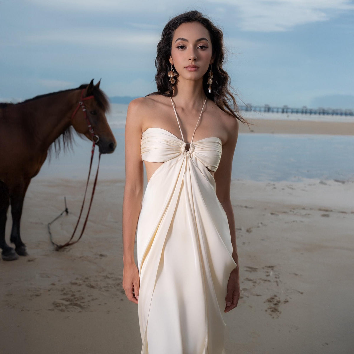 May include: A cream-colored, strapless dress with a halter neck and a cutout design. The dress is flowing and elegant, with a gathered bodice and a long skirt. The model is standing on a sandy beach with a horse in the background.