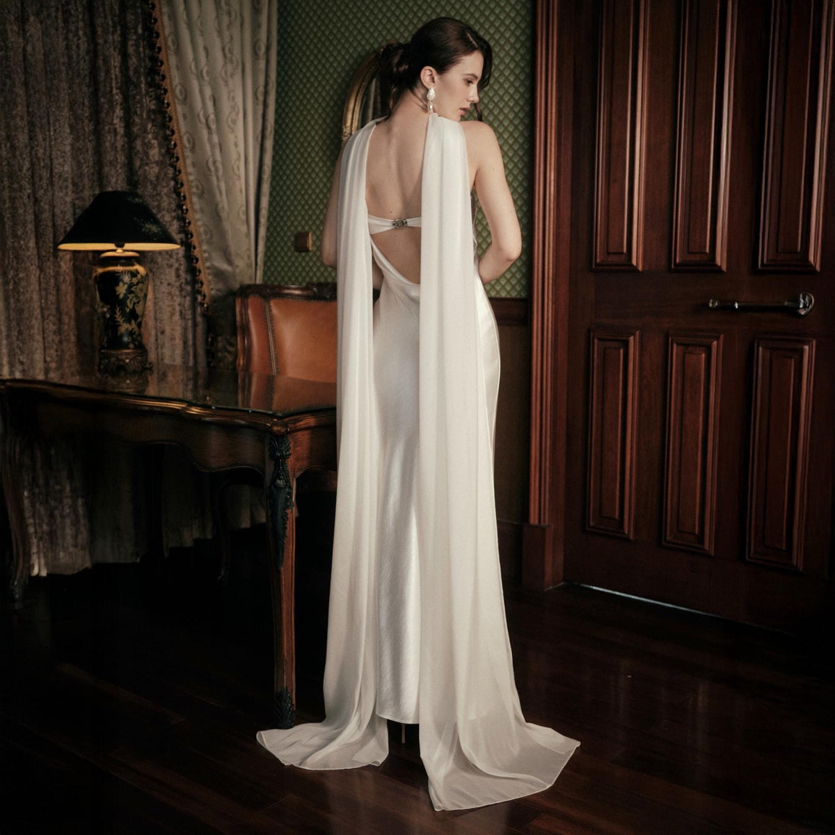May include: A white satin gown with a low back and flowing cape detail. The elegant dress features a simple silhouette and a long train. The model is standing in a room with dark wood furniture and a vintage lamp.