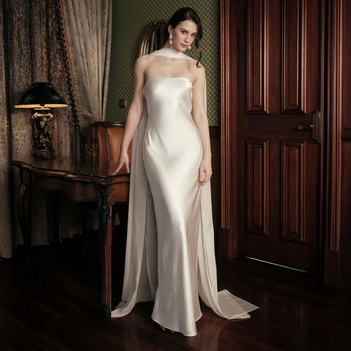 May include: A strapless, ivory-colored gown with a flowing train. The dress is made of a silky material and has a simple, elegant design. A matching scarf drapes over the shoulders. The setting is a room with dark wood furniture and a patterned wall.