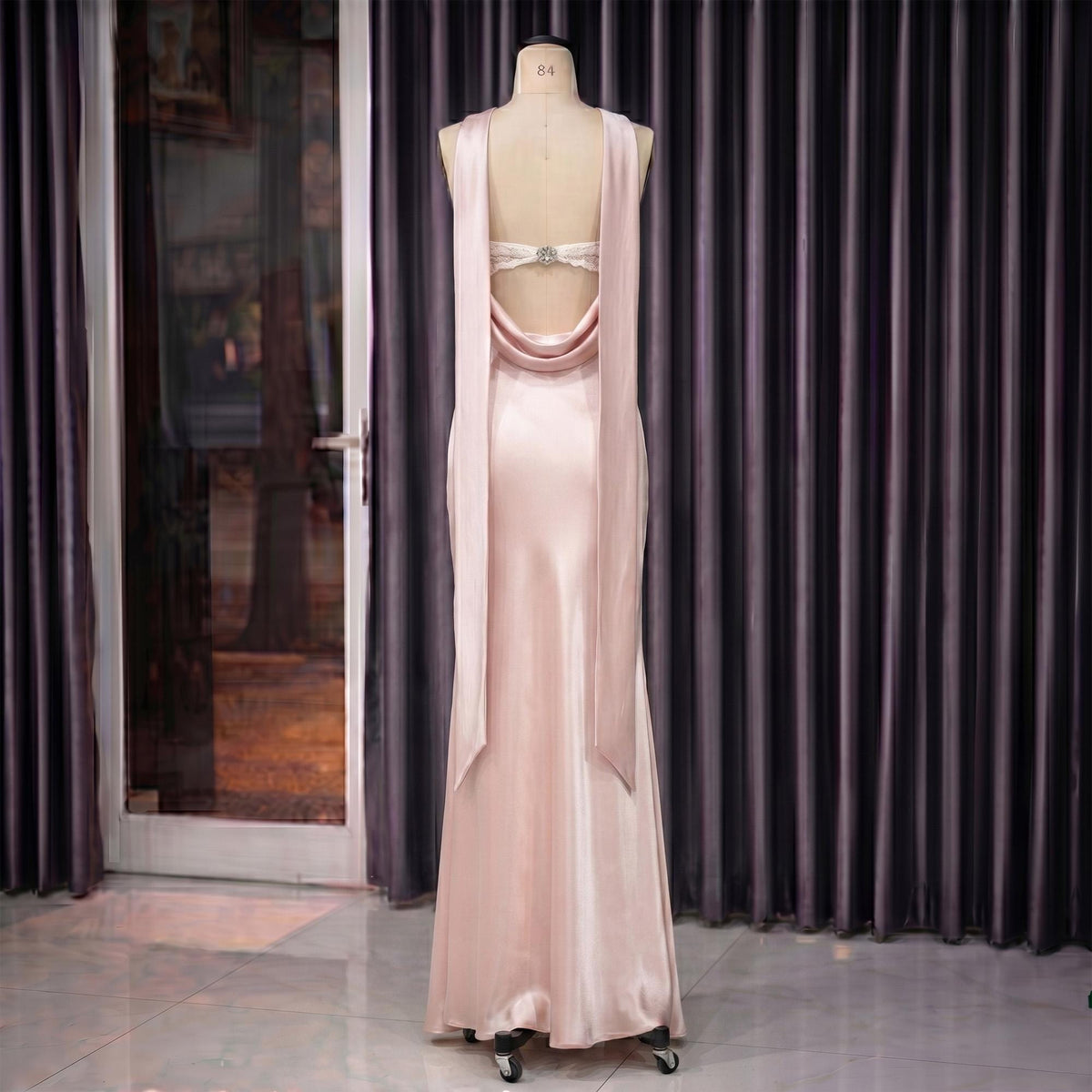 May include: A pale pink satin gown on a mannequin. The dress features a cowl neckline, open back, and long, flowing panels that drape down the back. A decorative embellishment is at the upper back. The dress is floor-length.
