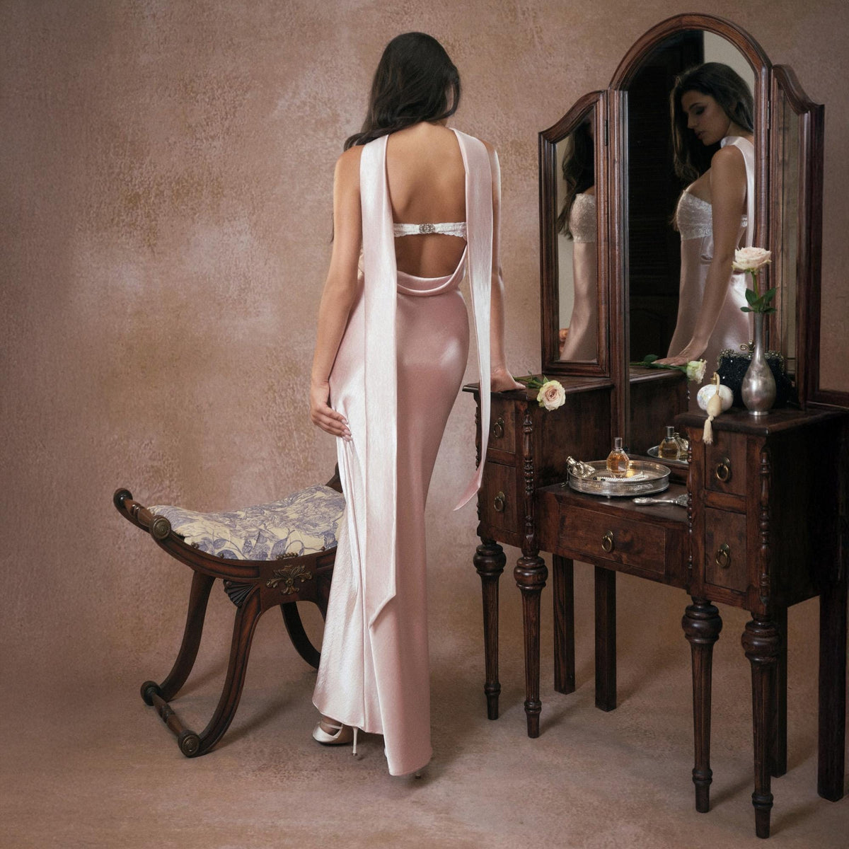 May include: A pale pink satin gown with a low back and long, flowing panels. The dress is worn with white shoes. A vintage vanity with a mirror and a small bench are in the background.