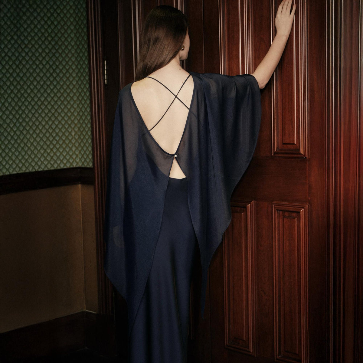 May include: A dark navy blue floor-length dress featuring a sheer, flowing cape-style overlay. The back design includes a deep scoop neckline with thin, crisscrossing straps and a single small button closure at the center of the upper back.