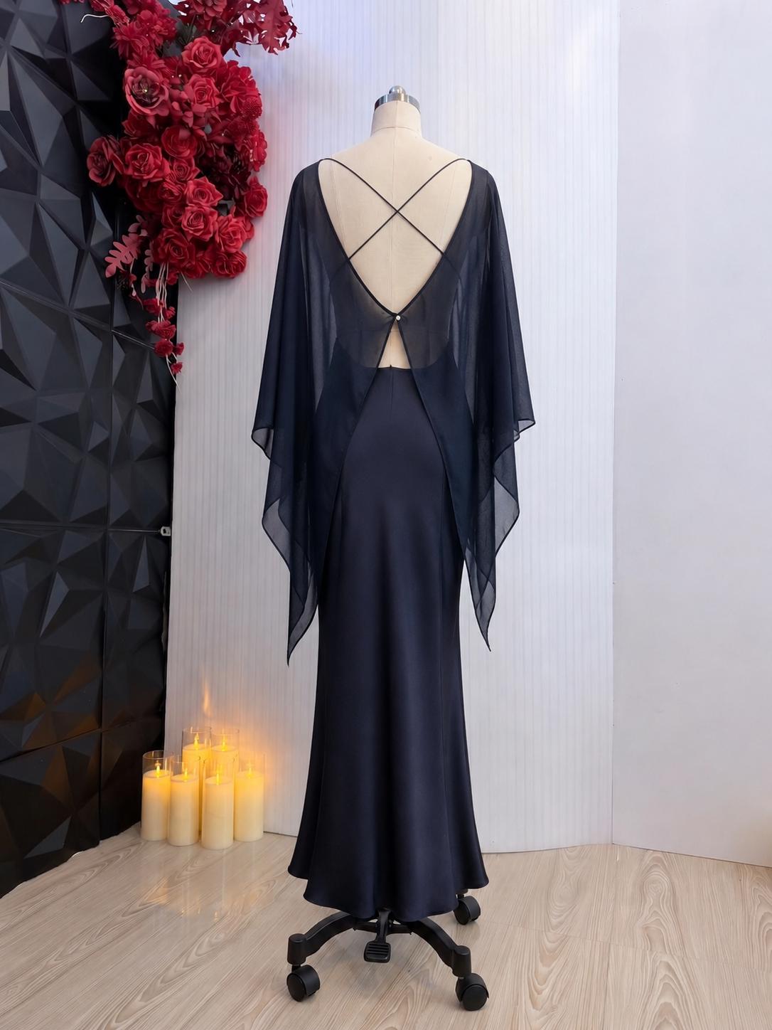 May include: A floor-length dark navy blue gown featuring a satin-finish skirt and a sheer, flowing cape-style overlay. The back displays a deep V-neckline with thin, crisscrossing straps and a single small button closure at the center back.