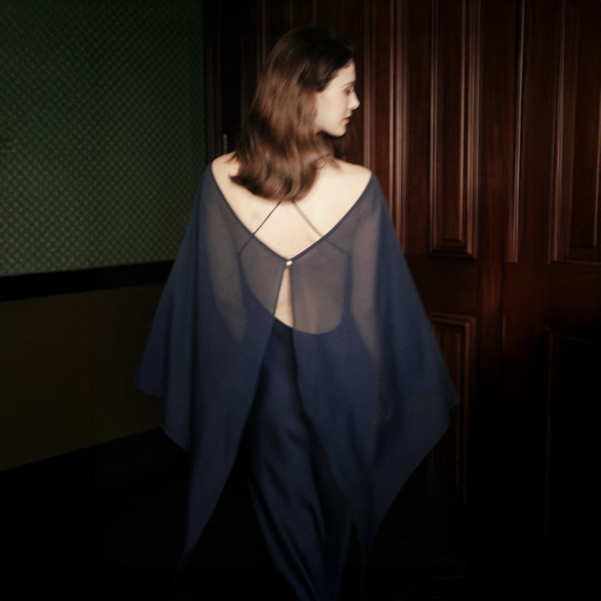 May include: A dark navy blue sleeveless dress featuring a sheer, flowing cape-like overlay that drapes over the shoulders and back. The garment includes thin, delicate straps that cross at the upper back, secured by a small, centered metallic button.